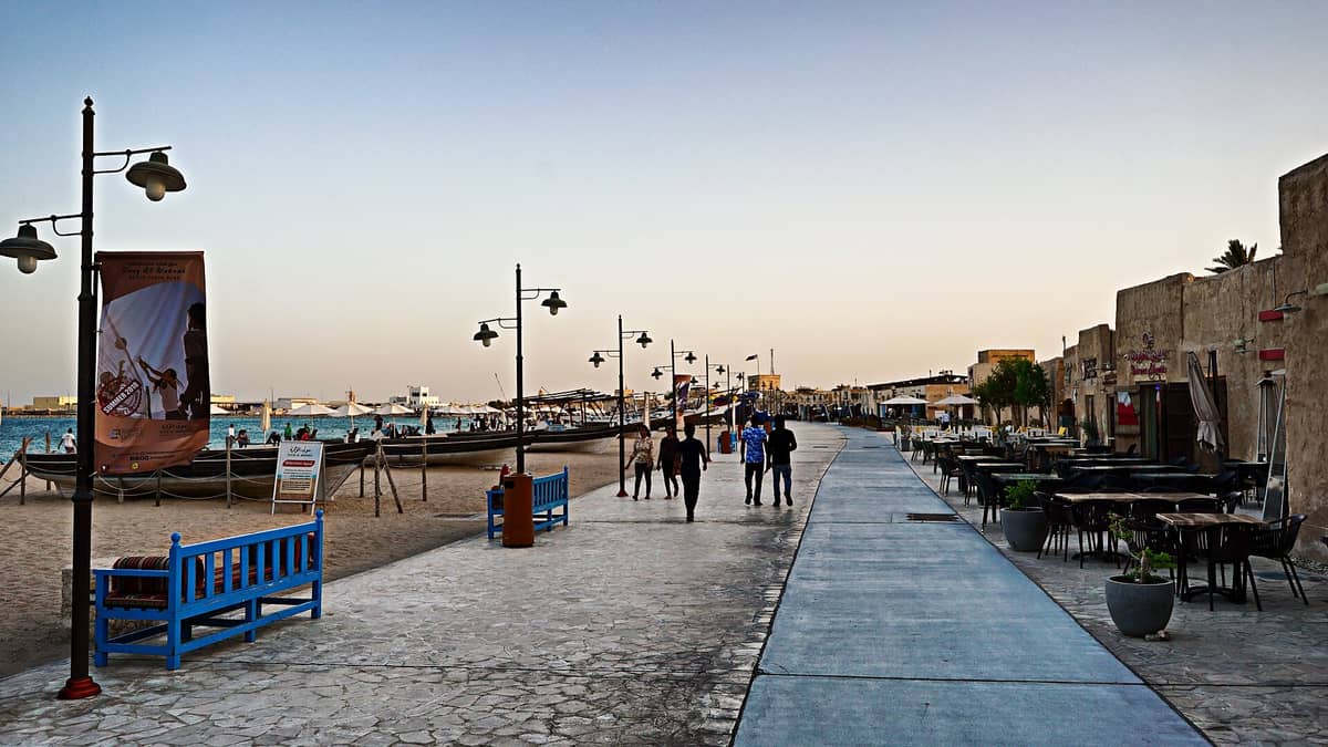 Die Al-Wakra-Corniche in Katar bei Sonnenuntergang: Menschen flanieren auf der Promenade, Boote liegen auf dem Sand, und traditionelle Souq-Gebäude mit Außensitzplätzen säumen das Ufer.