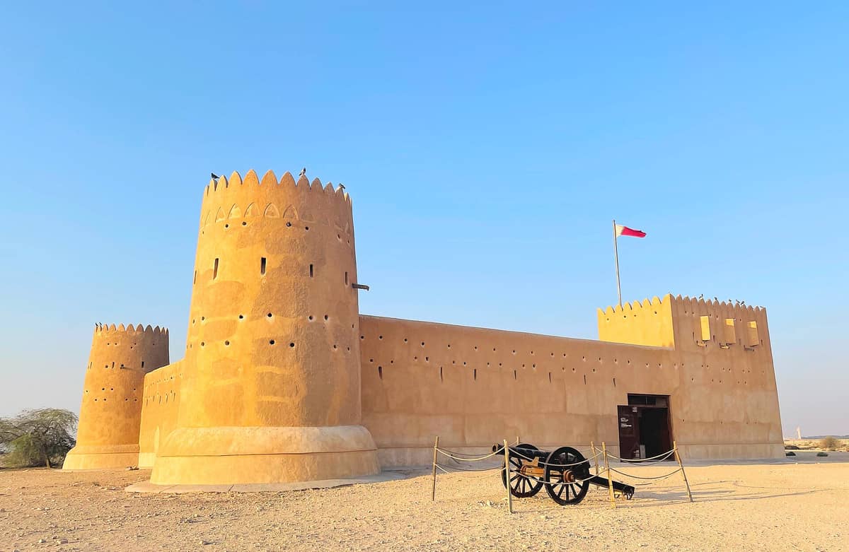 Weitwinkelpanorama des Al Zubarah Forts in Katar mit seinen markanten Rundtürmen, sandfarbenen Mauern, der katarischen Flagge und einer historischen Kanone im Vordergrund.