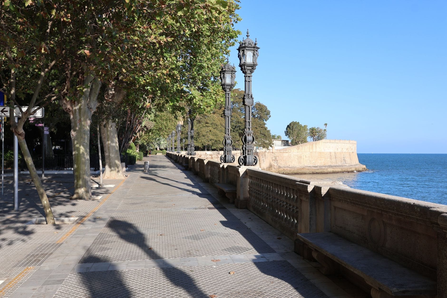 A wide promenade lined with trees, ornate street lamps, stone benches, and a stone balustrade overlooking the blue waters of the Bay of Cádiz.