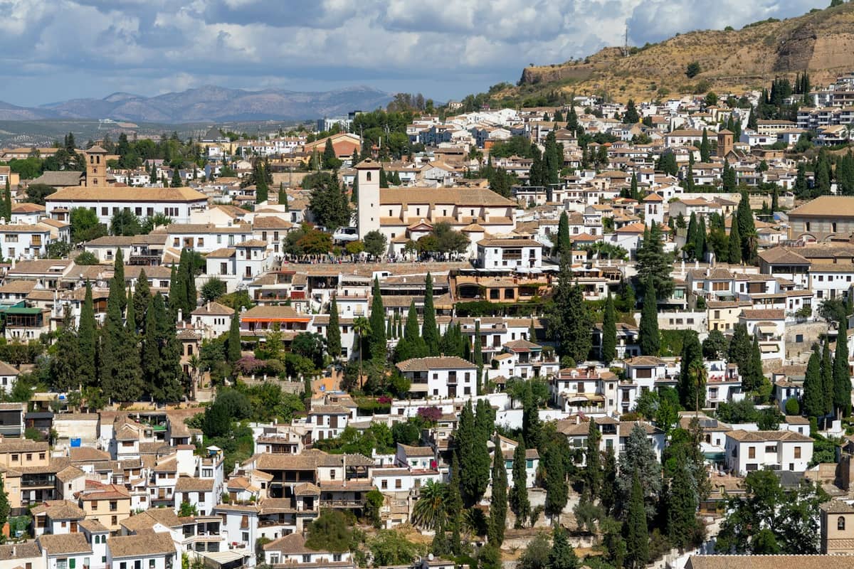 วิวพาโนรามาของย่าน Albaicín ในเมือง Granada พร้อมด้วยบ้านทาสีขาว ถนนแคบ และเนินเขาภายใต้ท้องฟ้าที่มีเมฆบางส่วน