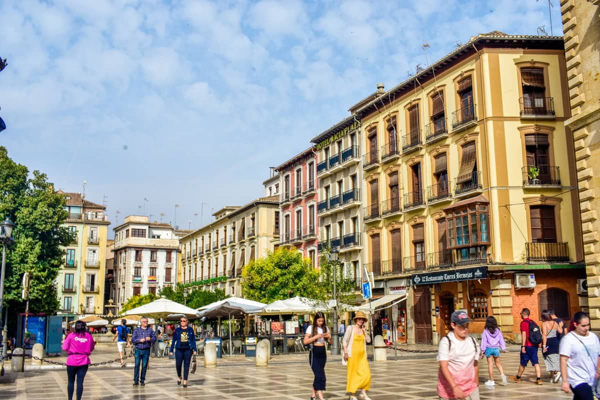 ผู้คนกำลังเดินและพักผ่อนที่ Plaza Nueva จัตุรัสกลางเมืองที่มีอาคารประวัติศาสตร์สีสันสดใสและคาเฟ่กลางแจ้งในเมืองกรานาดา