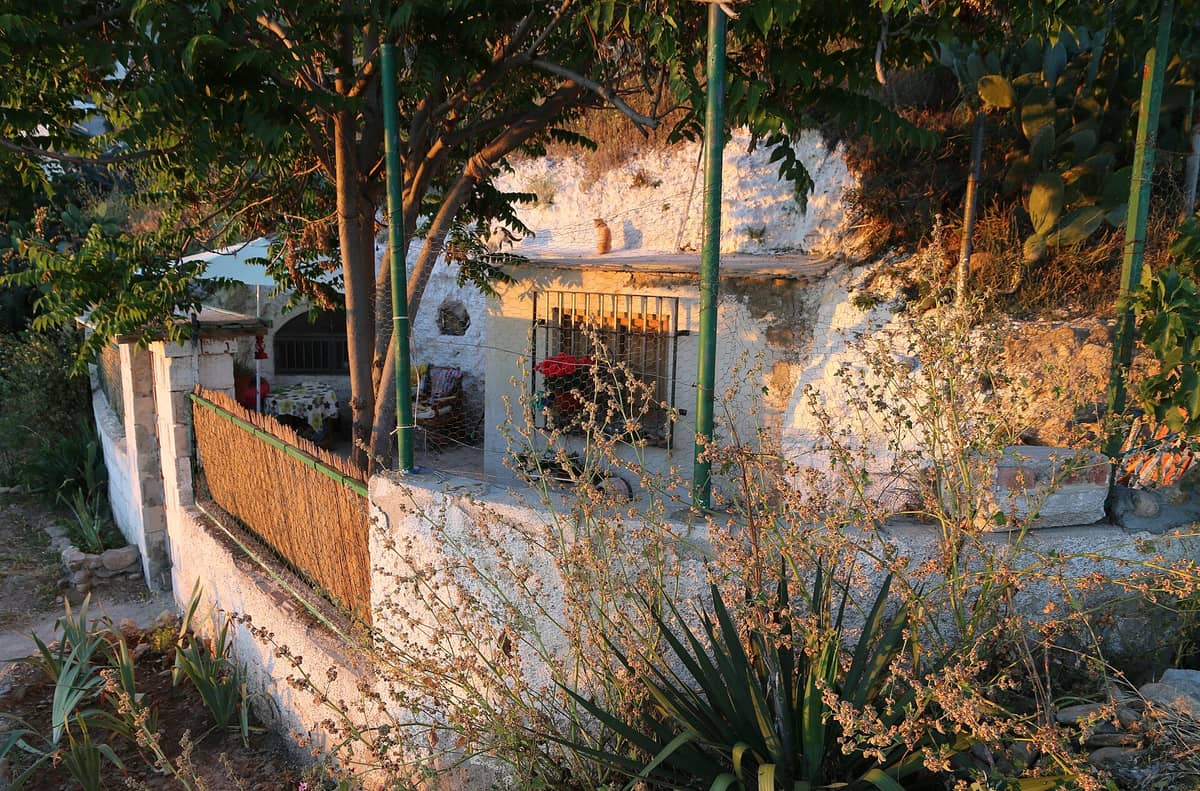 บ้านถ้ำสีขาวสดใสในแสงแดดที่ Sacromonte เมือง Granada พร้อมต้นไม้และพืชพรรณในเบื้องหน้าและเนินเขาเป็นฉากหลัง