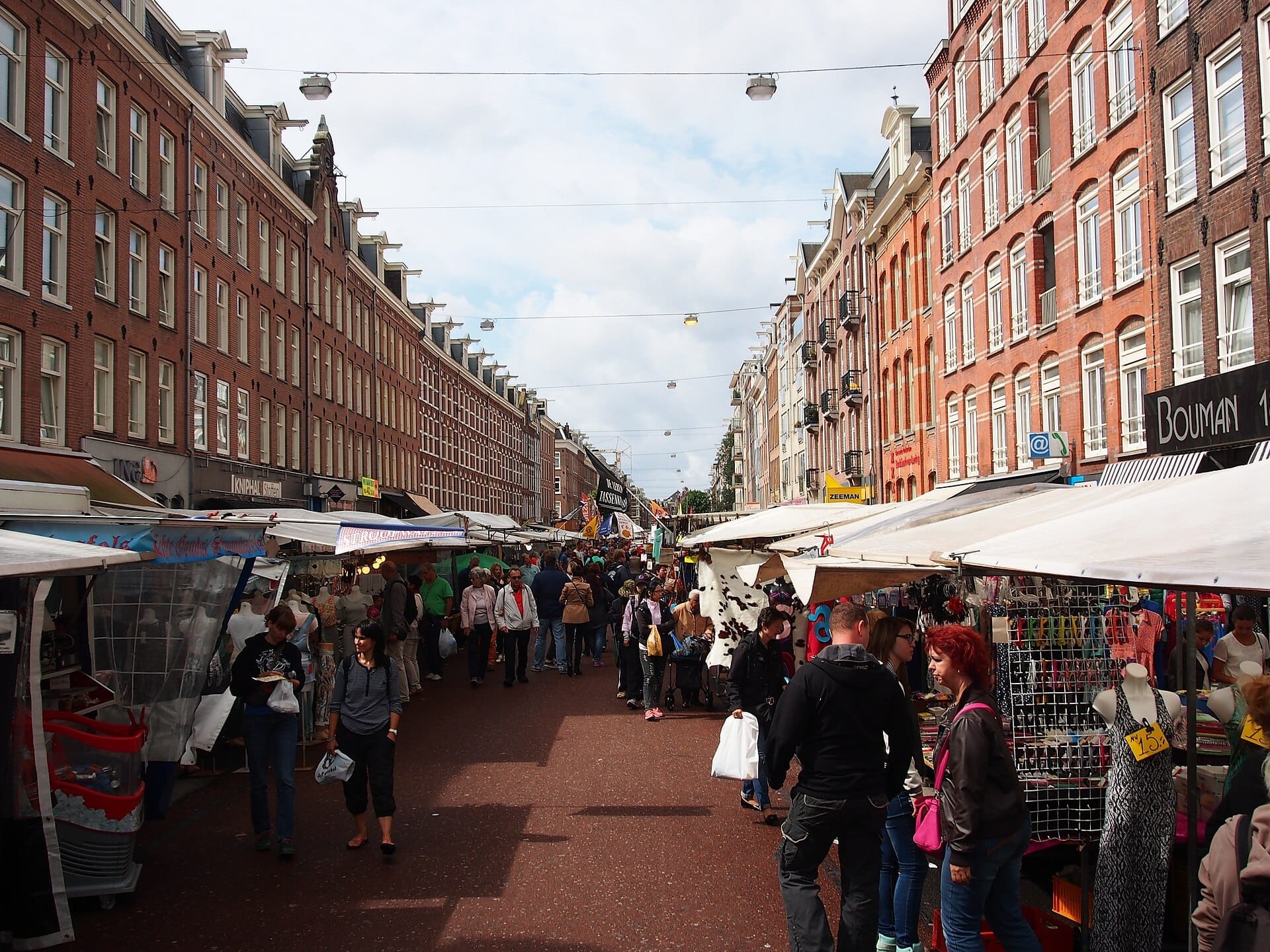 Compradores exploran puestos y productos coloridos en el Mercado Albert Cuyp de Ámsterdam, flanqueado por históricos edificios de ladrillo rojo bajo un cielo despejado.