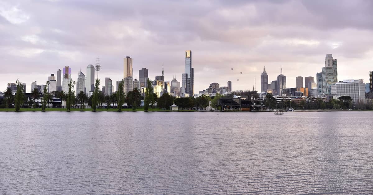 Vista panorámica de Albert Park Lake con el agua en calma en primer plano, palmeras y el skyline de Melbourne recortándose bajo un cielo nublado.