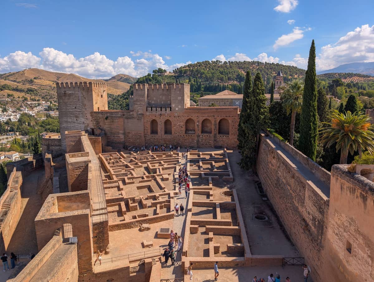 ภาพวิวแนวกว้างของ Alcazaba ในอัลฮัมบร้า กำแพงป้อม หอคอยป้องกัน เหล่านักท่องเที่ยวเดินท่ามกลางซากโบราณภายใต้ท้องฟ้าสีฟ้าใส
