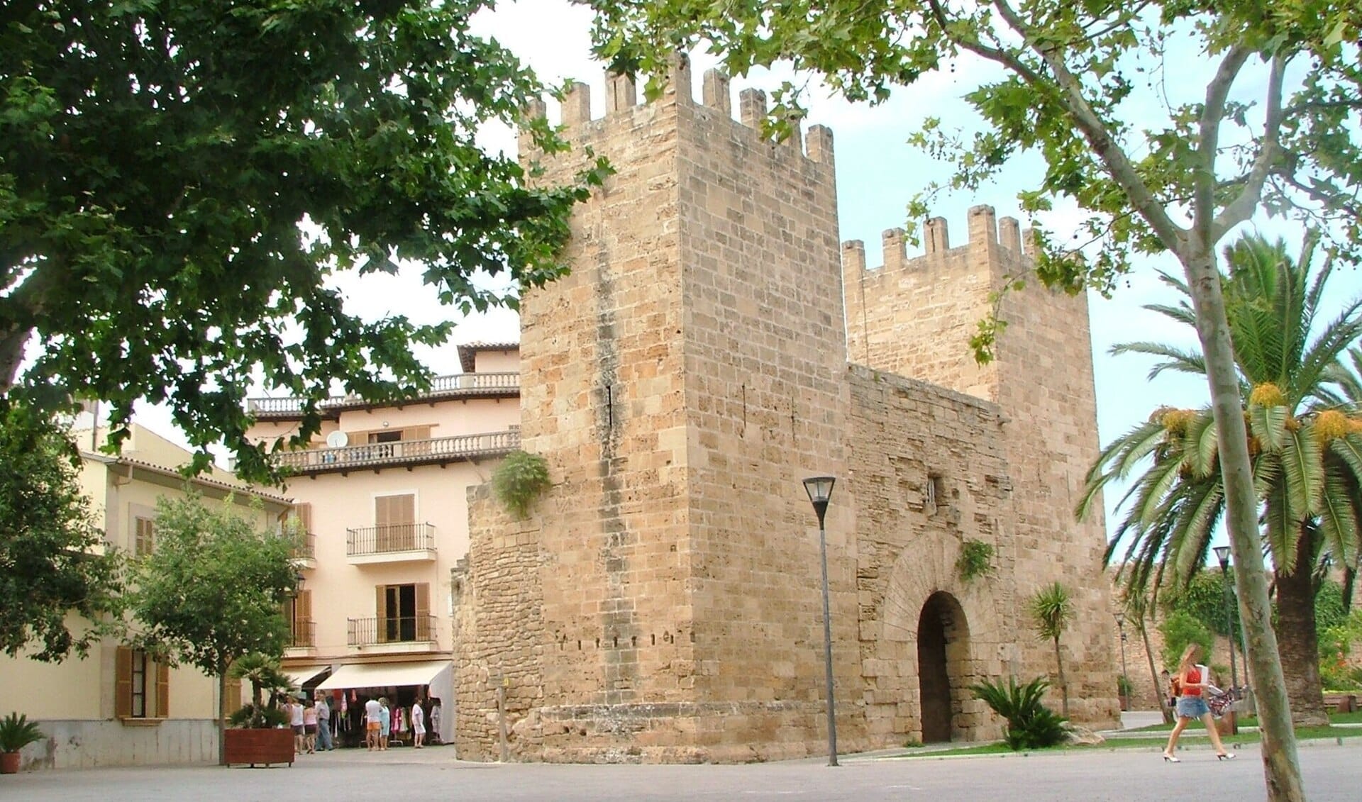 Mura medievali in pietra e torri del centro storico di Alcúdia, Maiorca, circondate da alberi frondosi e vicini edifici storici durante il giorno.