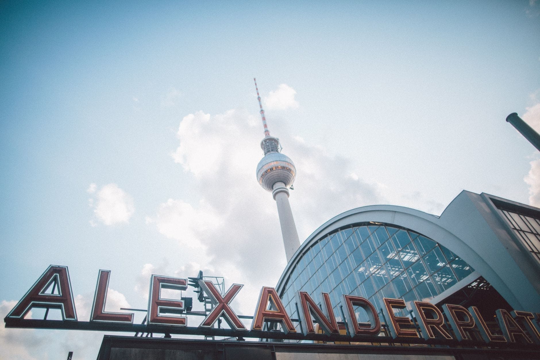Vista di Alexanderplatz con l'iconica Torre della Televisione, la copertura in vetro curva della stazione, l'insegna al neon rossa di Alexanderplatz e un cielo luminoso sullo sfondo.