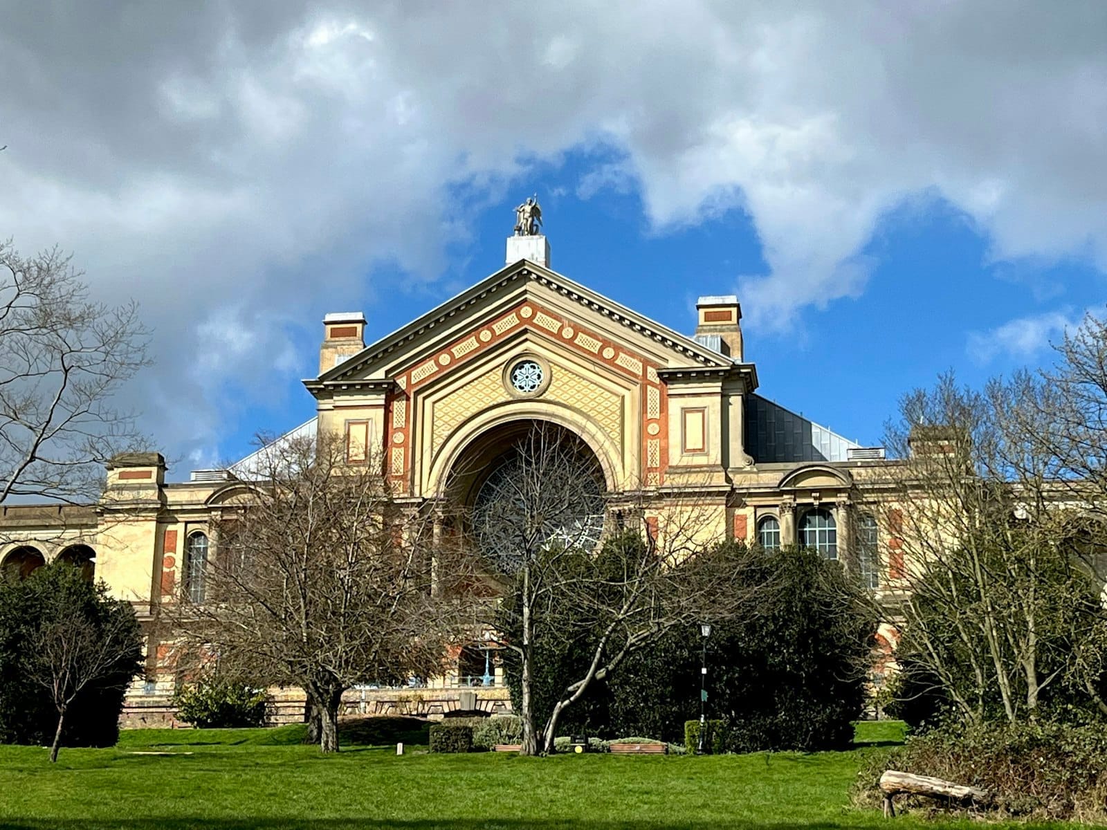 La sontuosa facciata vittoriana dell'Alexandra Palace si eleva sopra un parco verde, incorniciata dagli alberi e stagliatasi su un cielo azzurro brillante con nuvole spettacolari.
