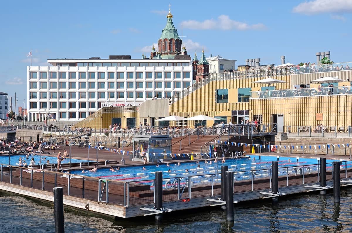 Allas Sea Pool em Helsinque com piscinas em uma plataforma flutuante, pessoas nadando e tomando sol, prédios modernos e a Catedral Uspenski ao fundo.
