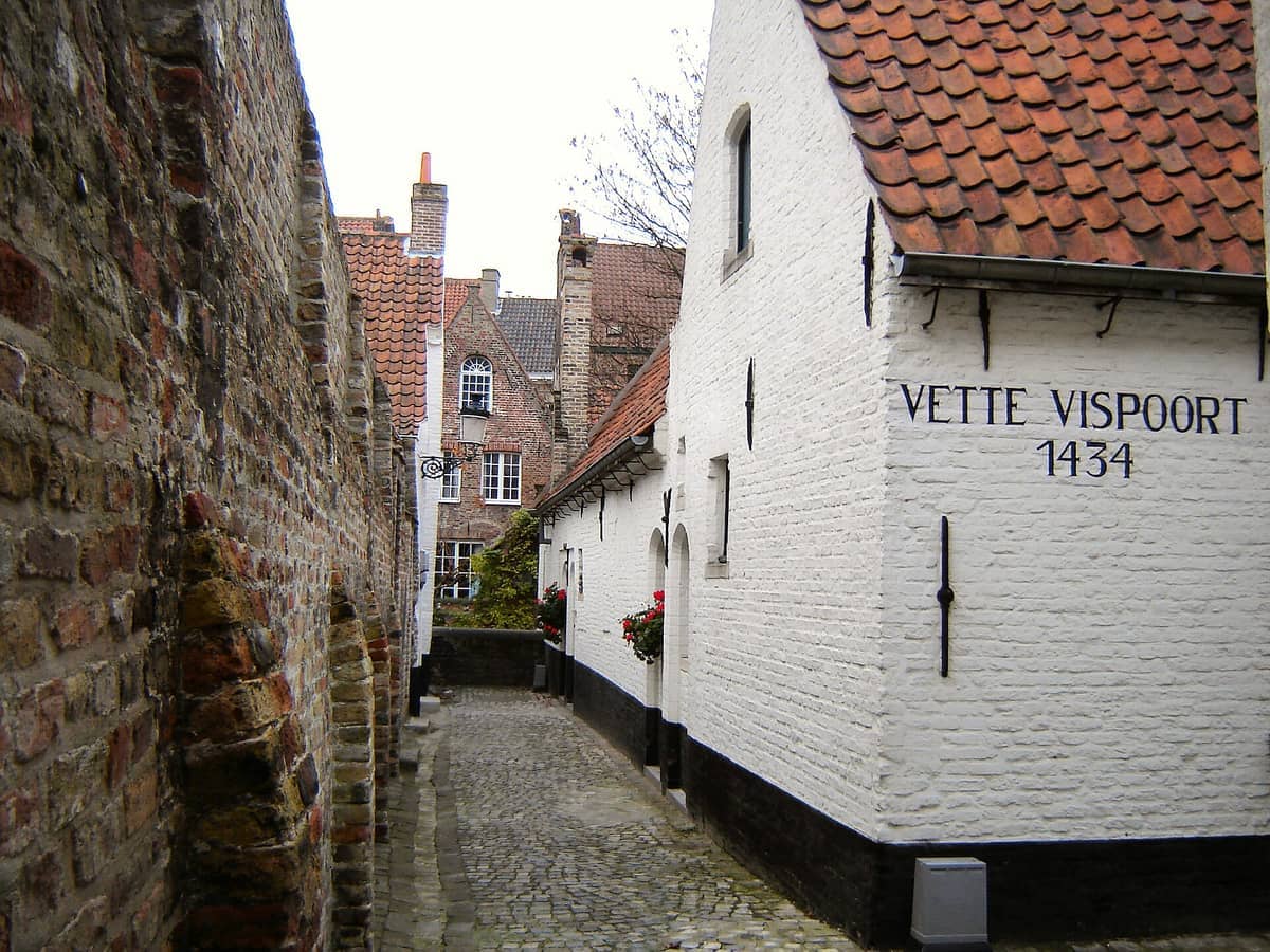 Historic Bruges almshouse courtyard showing whitewashed brick buildings with red roofs, cobblestone path, and the sign 'Vette Vispoort 1434' on the wall.