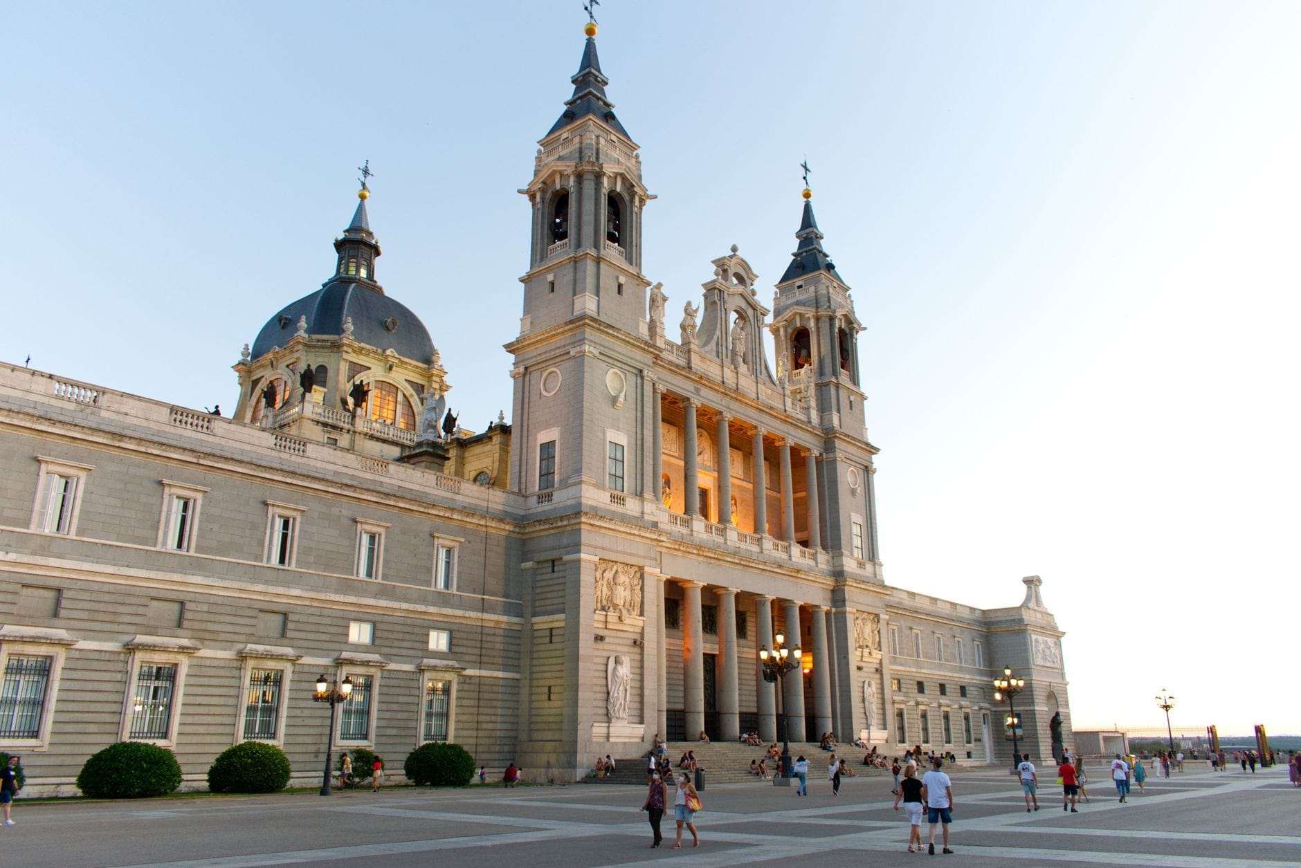 La Cattedrale dell'Almudena al tramonto, avvolta da una luce calda, con la facciata e le torri in primo piano, visitatori sulla piazza e un cielo terso di sera.