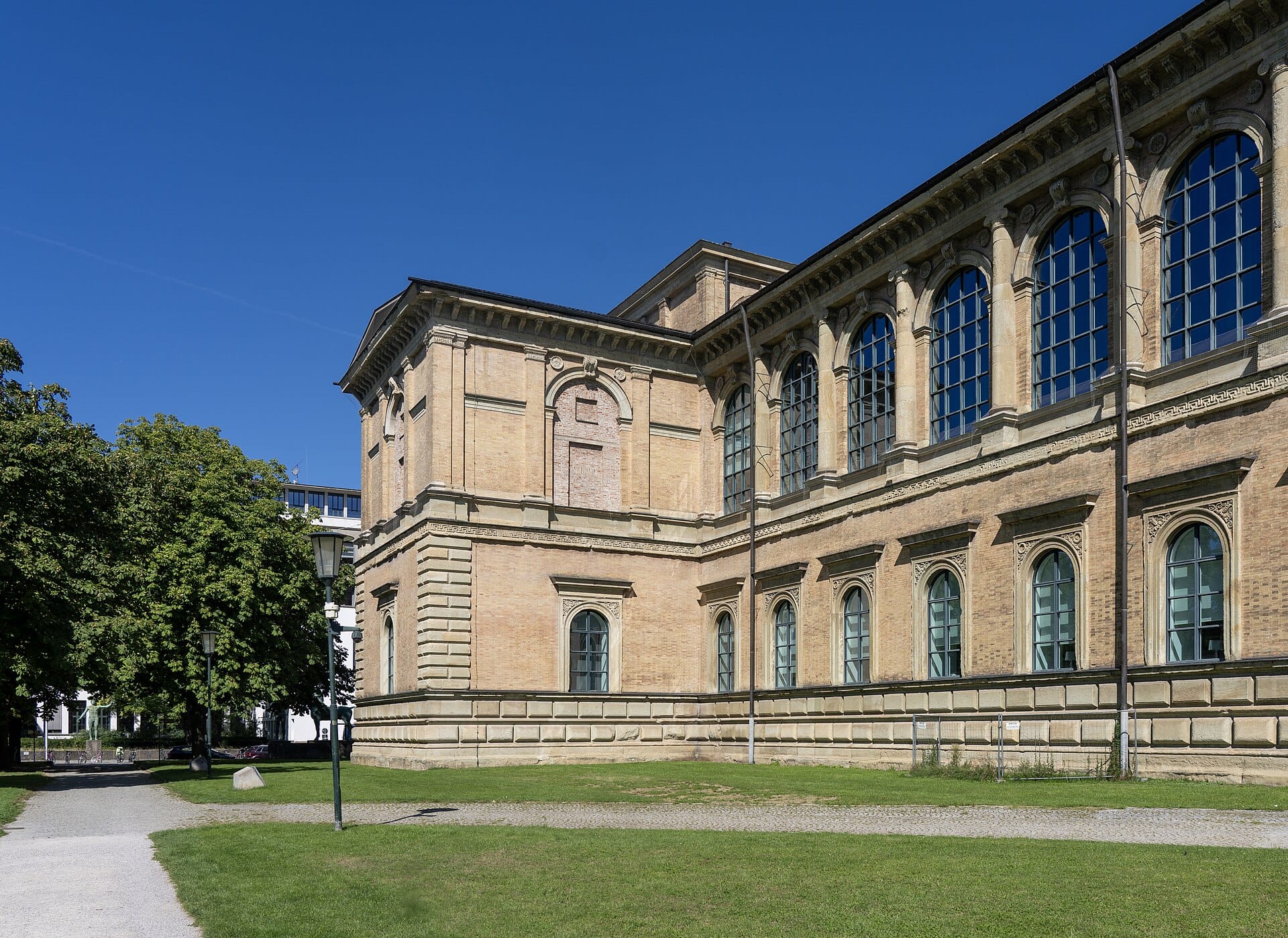 มุมมองด้านนอกของ Alte Pinakothek ในมิวนิก เห็นหน้าต่างโค้งอันเป็นเอกลักษณ์ ผนังอิฐเหลือง สนามหญ้าเขียวขจีใต้ท้องฟ้าสีฟ้าใส