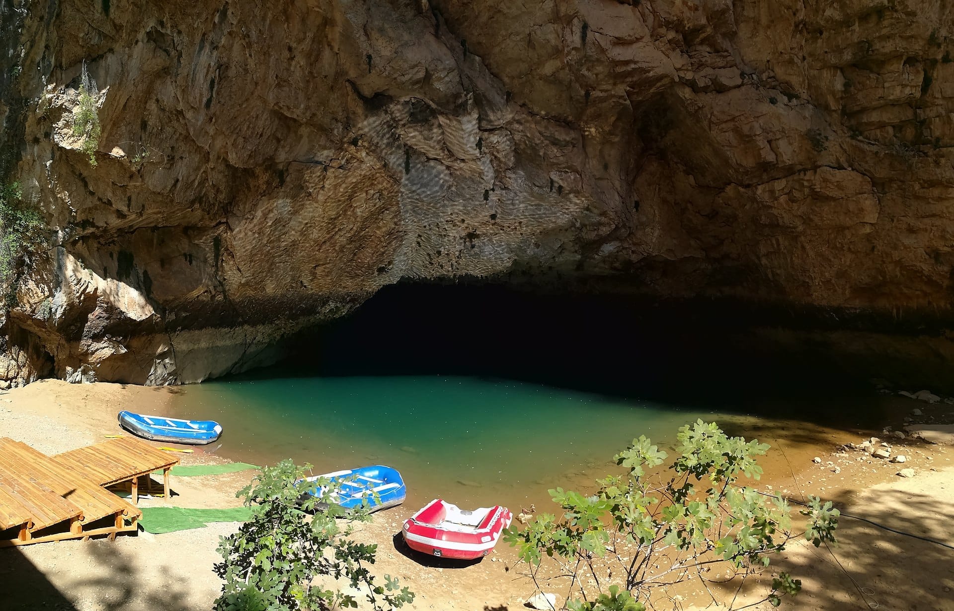 Ingresso della Grotta Altınbeşik con lago sotterraneo turchese, pareti rocciose frastagliate e diversi gommoni colorati sulla riva sabbiosa.