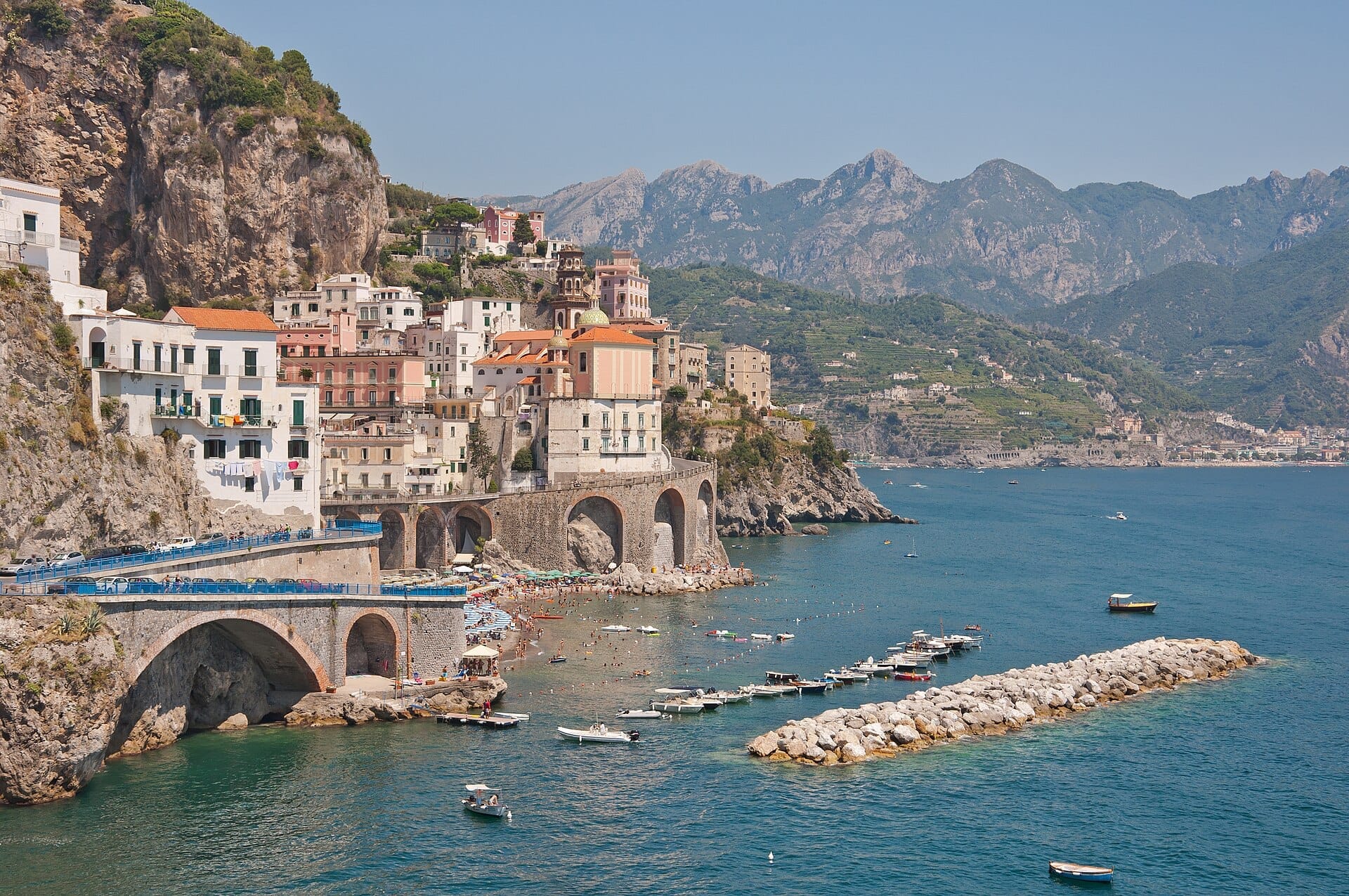 Le case colorate di Atrani sulla Costiera Amalfitana affacciate sul mare azzurro, con barche sull'acqua e montagne sullo sfondo.