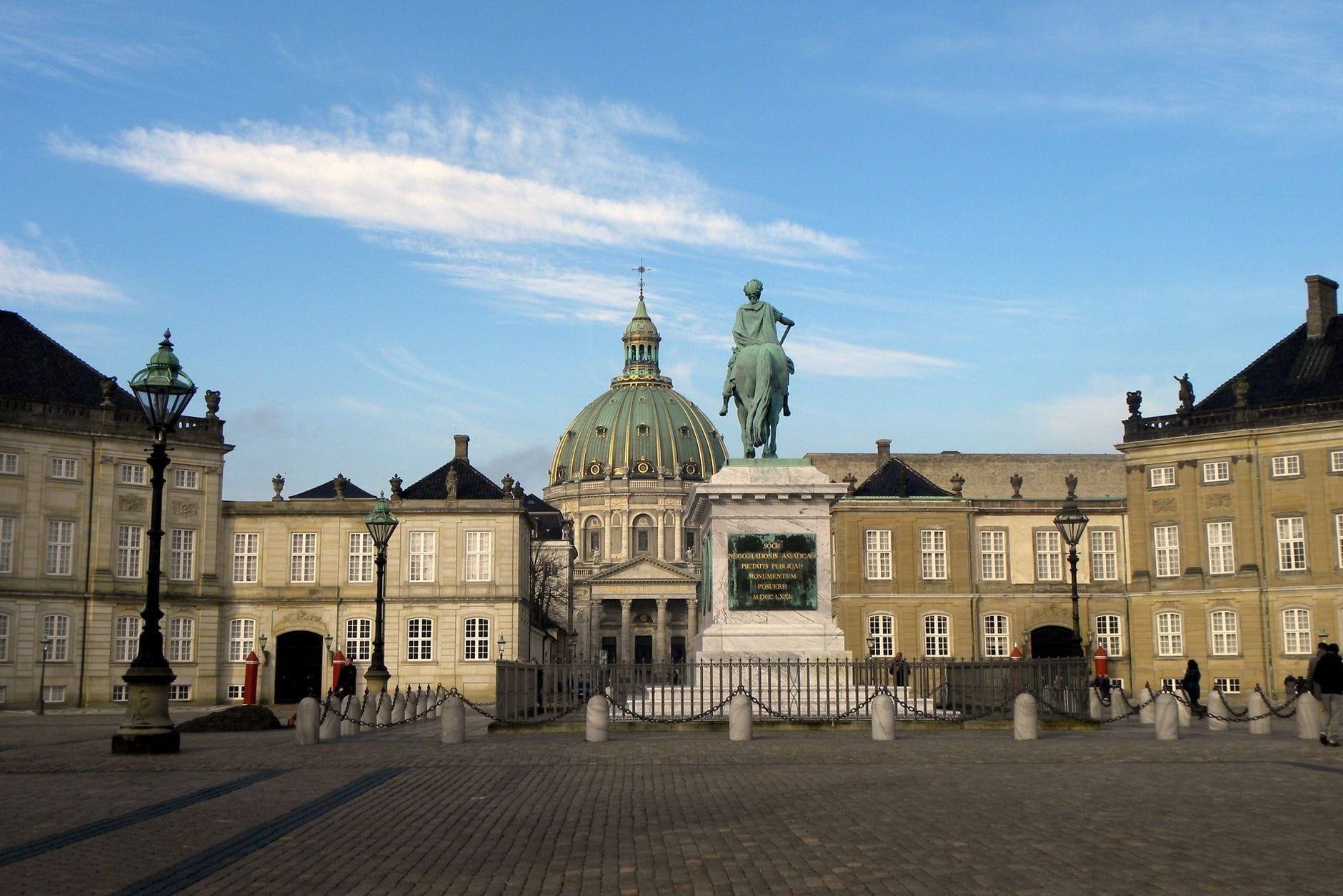 Vista panoramica della piazza di Amalienborg con la statua equestre centrale e la cupola della Chiesa di Frederik sullo sfondo, sotto un cielo azzurro limpido.
