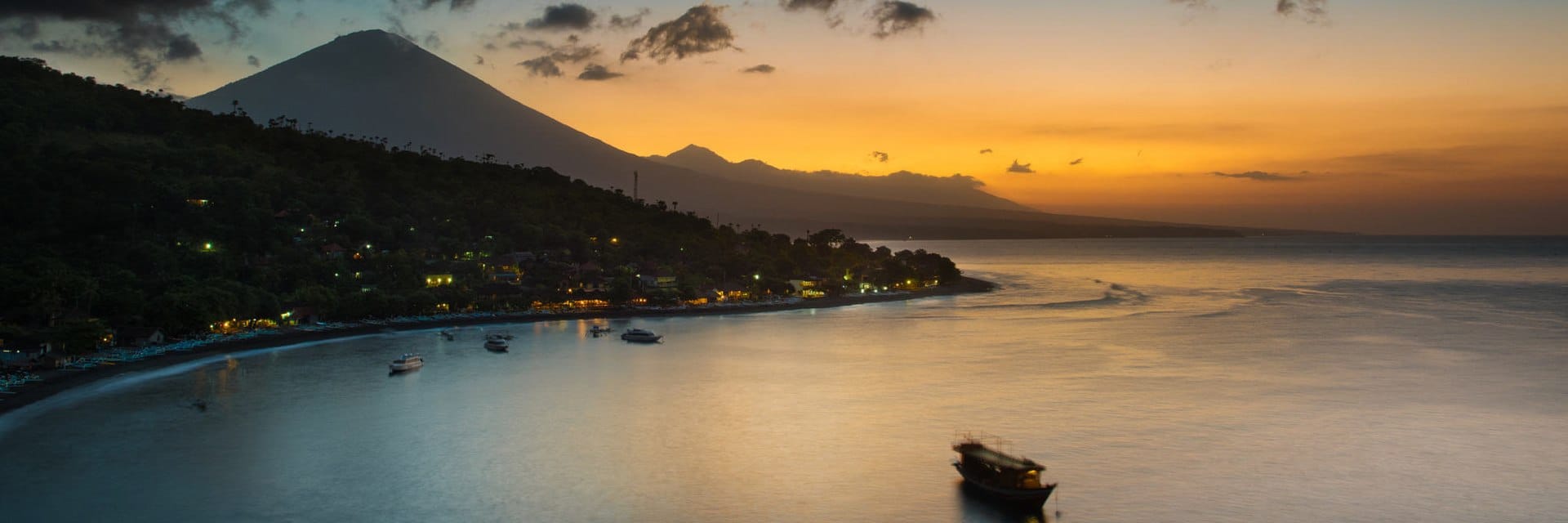 Tramonto tranquillo su Amed, Bali, con riflessi dorati sulle rive di ciottoli, barche da pesca e la maestosa sagoma del Monte Agung.
