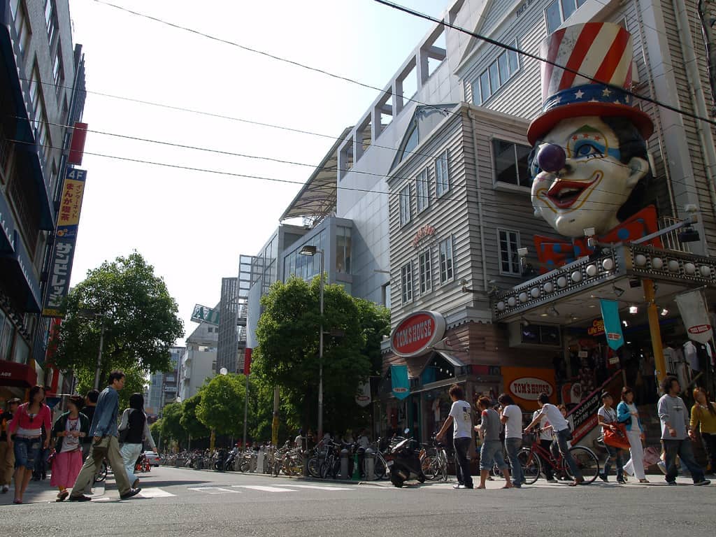 Pedestrians cross a busy street in Amerikamura, Osaka, with a large clown-faced shop sign and modern buildings under bright daylight.