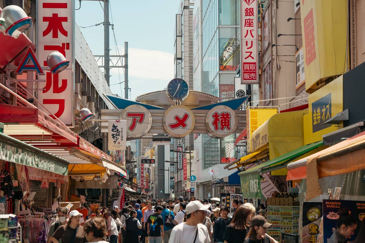 Bustling Ameyoko Market street view in Tokyo with crowds of shoppers, iconic 'Ameyoko' sign, colorful store awnings, and vibrant atmosphere on a sunny day.