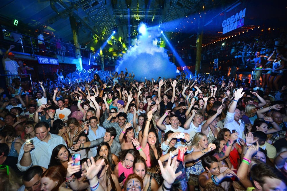 Grande folla che balla sotto luci colorate e nebbia all'interno della discoteca Amnesia Ibiza, con mani alzate e vibrante atmosfera di festa.