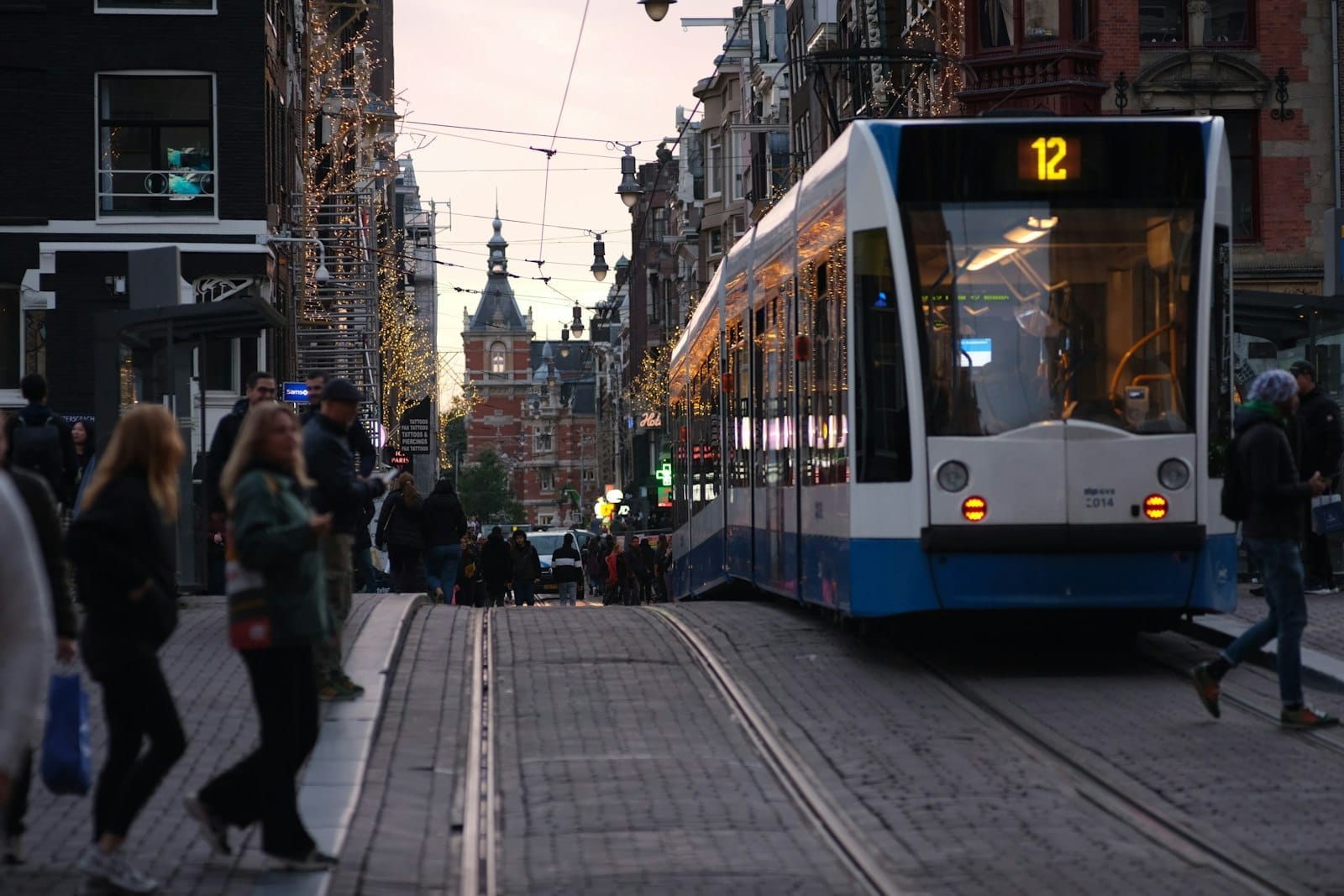 Một chiếc tàu điện màu xanh và trắng di chuyển trên con phố nhộn nhịp ở Amsterdam với người đi bộ và các tòa nhà Hà Lan truyền thống ở phía sau.