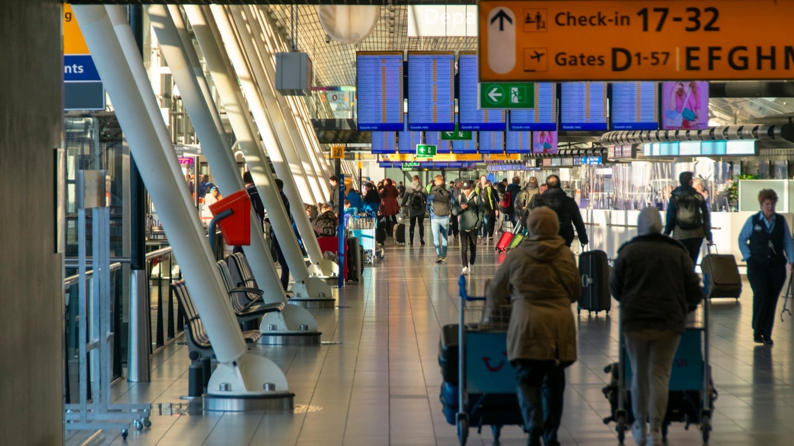 Terminal concurrida del Aeropuerto de Amsterdam Schiphol con viajeros, carritos de equipaje, pantallas de salidas y señalización amarilla bajo una brillante iluminación interior.
