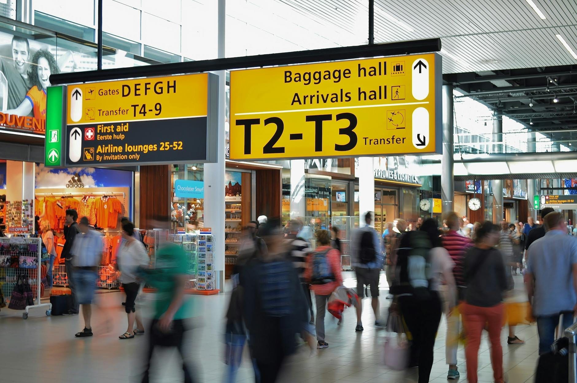 Concurrida escena interior en el Aeropuerto de Schiphol con tiendas, cafeterías y personas caminando bajo letreros amarillos brillantes de la terminal.