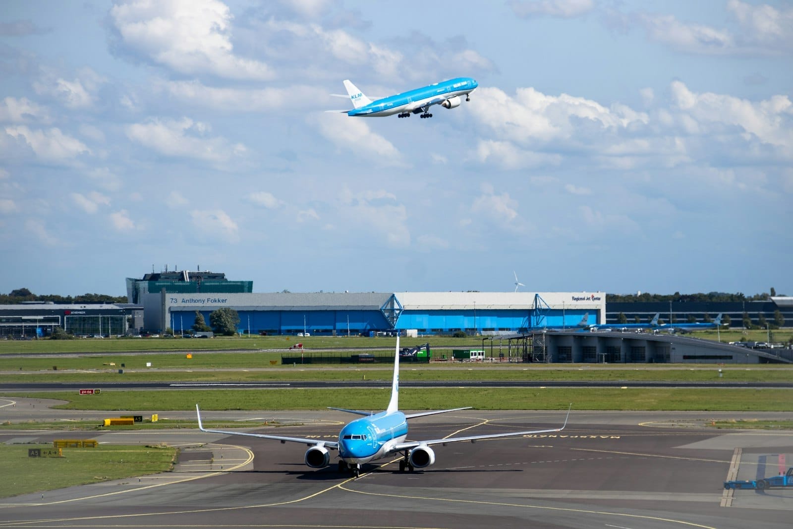 Aviones de KLM en la pista y despegando en el Aeropuerto de Amsterdam Schiphol, con cielos azules y edificios del aeropuerto al fondo.