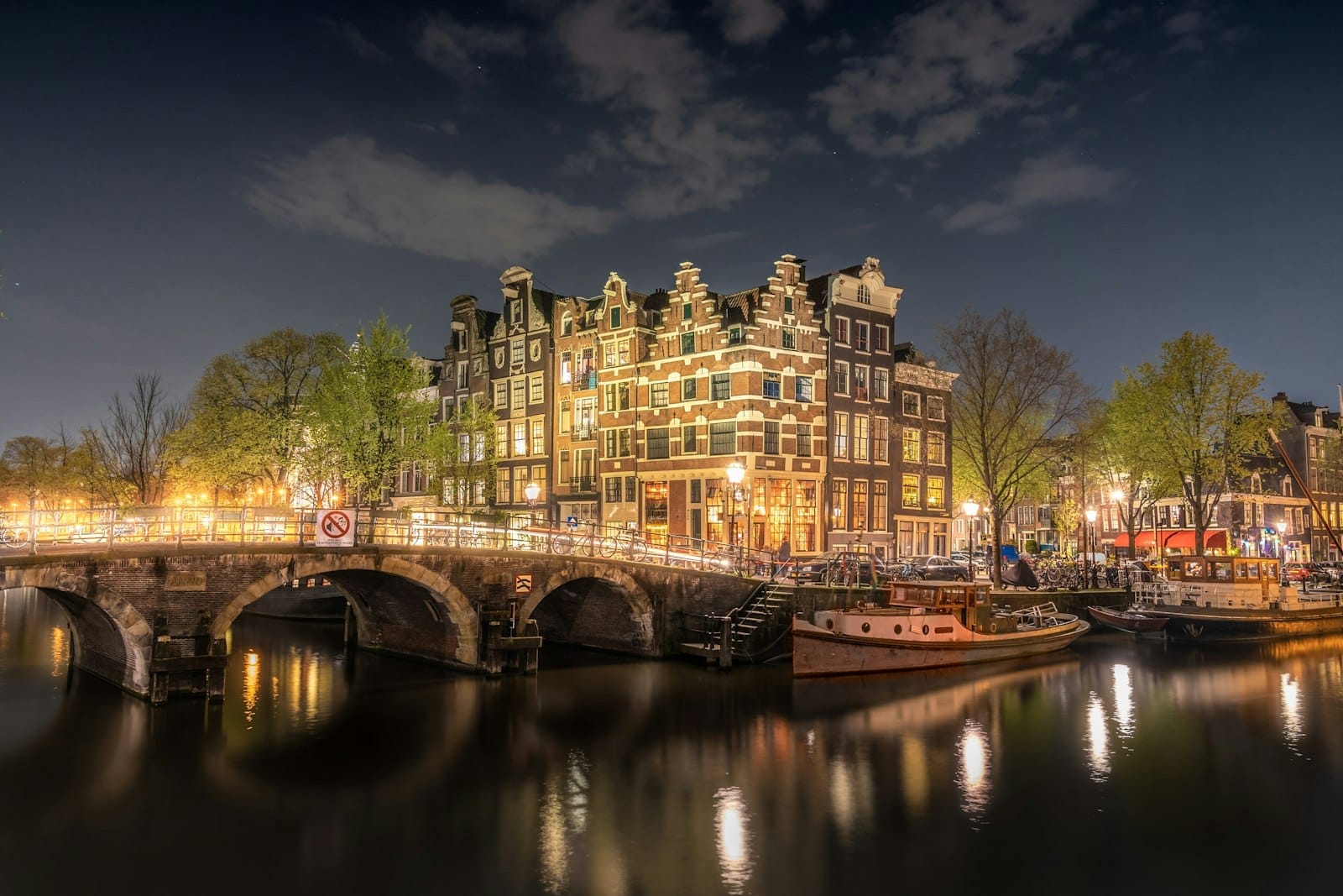 Vista nocturna de las iluminadas casas del canal de Ámsterdam y un puente arqueado reflejado en el agua, con barcas y árboles bajo un cielo dramático.
