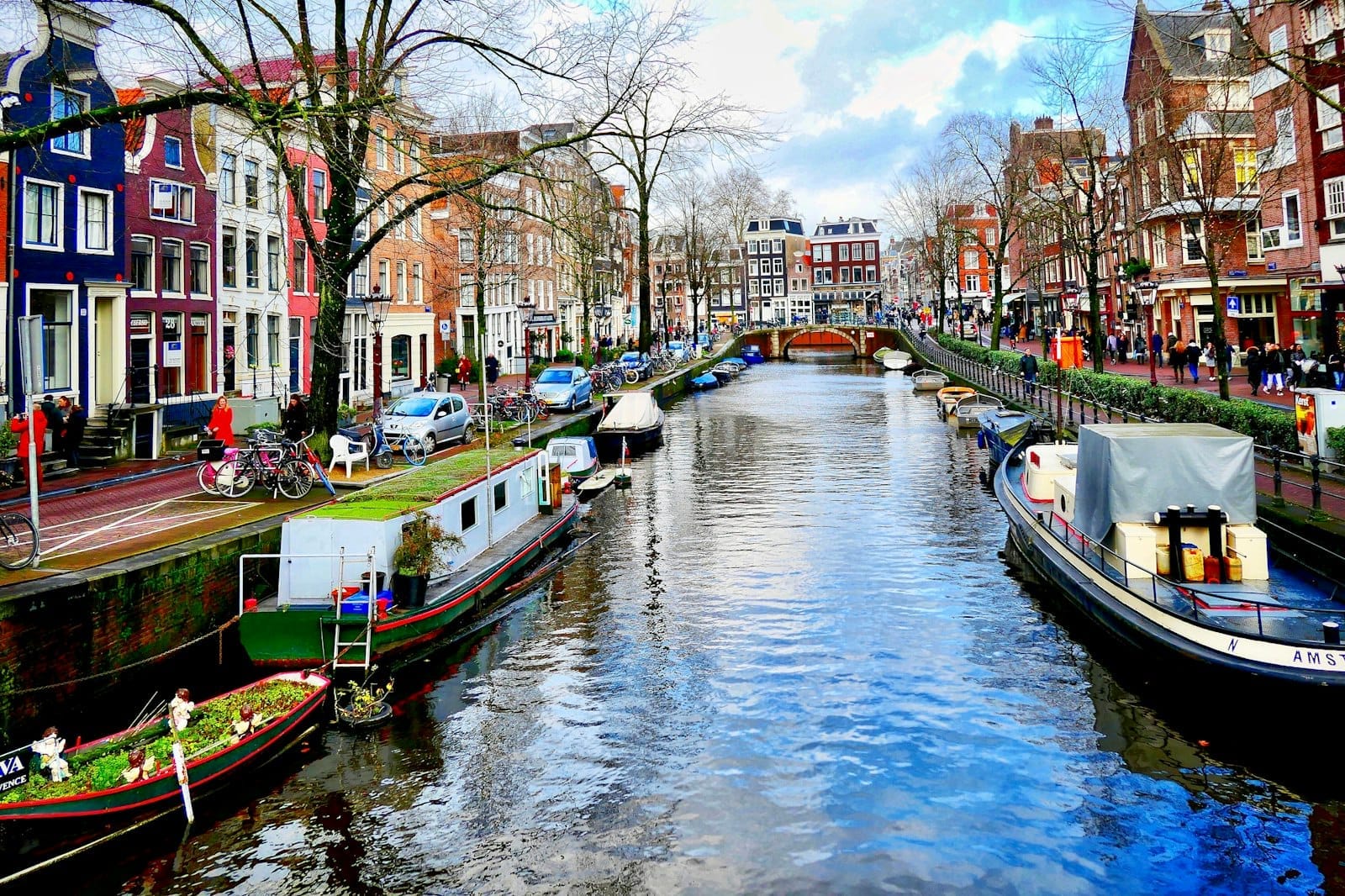 Cảnh kênh đào Amsterdam rực rỡ với những ngôi nhà hẹp truyền thống, thuyền bè và kênh đào rợp bóng cây dưới bầu trời xanh.