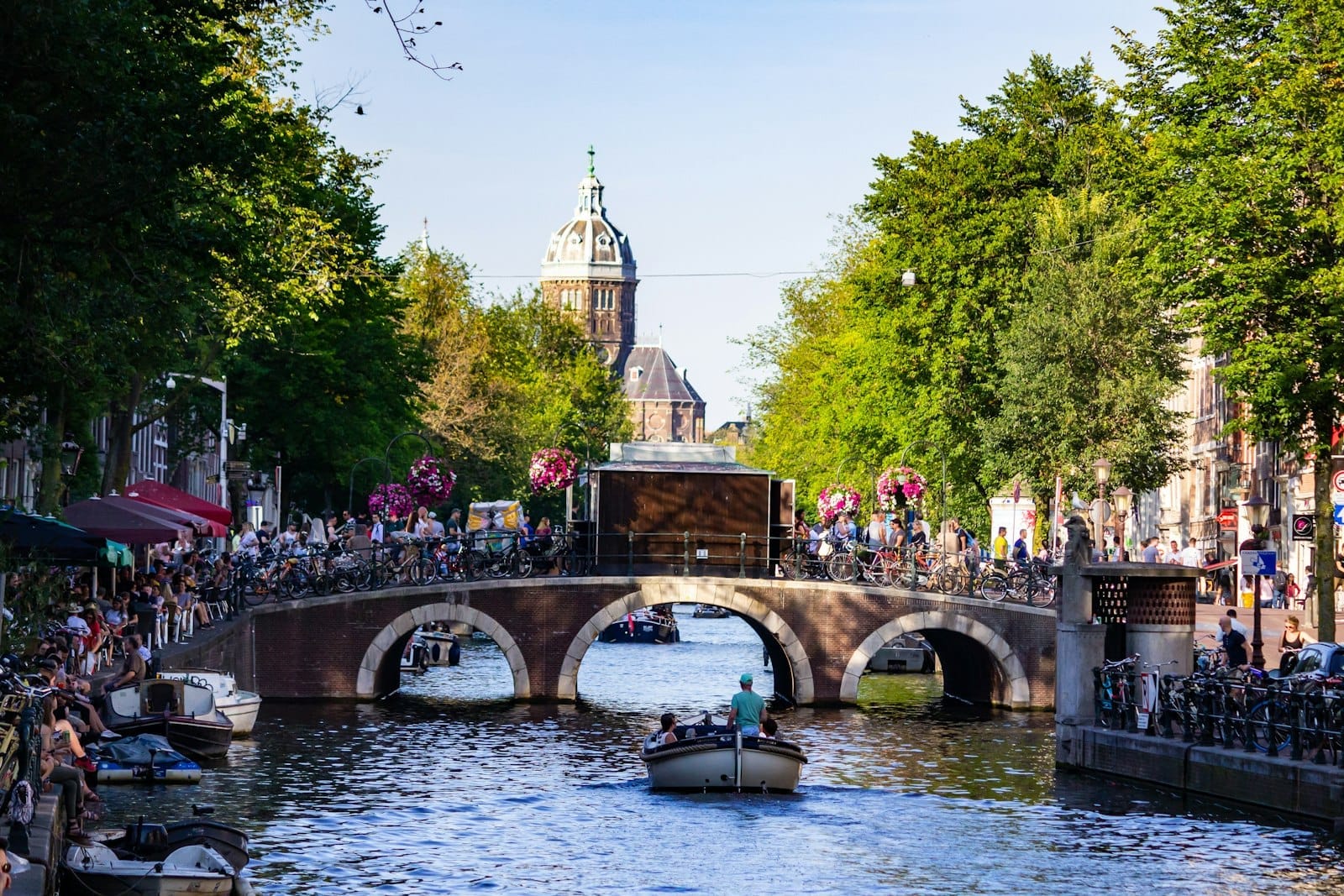 Kênh đào Amsterdam nhộn nhịp với thuyền bè, một cây cầu đá đông người qua lại, cây cối xanh tươi và các tòa nhà lịch sử dưới ánh nắng cuối mùa xuân hoặc mùa hè.