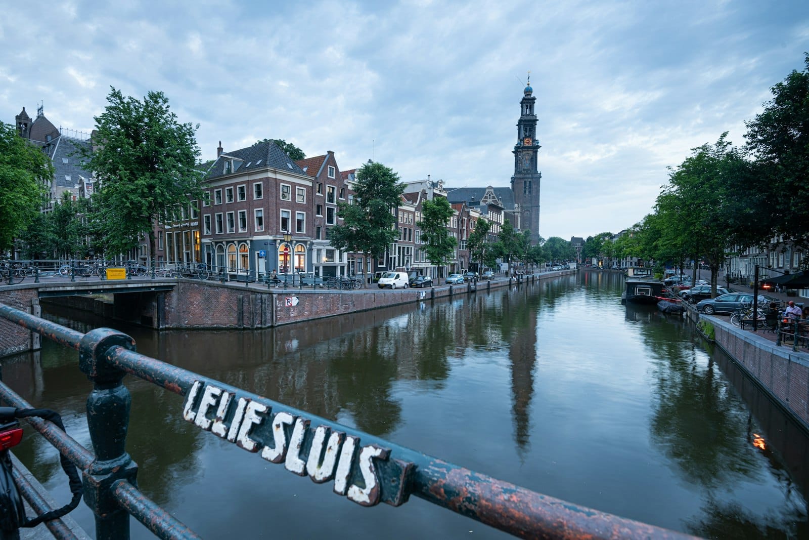 Vista clásica de un canal de Ámsterdam con casas históricas, árboles a orillas del canal y la torre de la iglesia Westerkerk, vista desde un puente con un cartel que dice 'Lelie Sluis'.