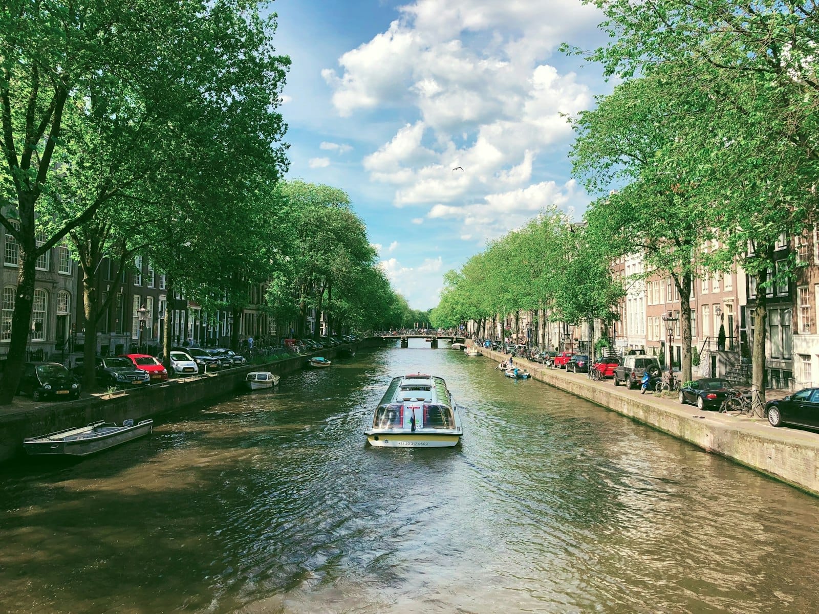 Un pintoresco canal de Ámsterdam con árboles a ambos lados, coches aparcados junto al agua y un barco navegando por el centro.