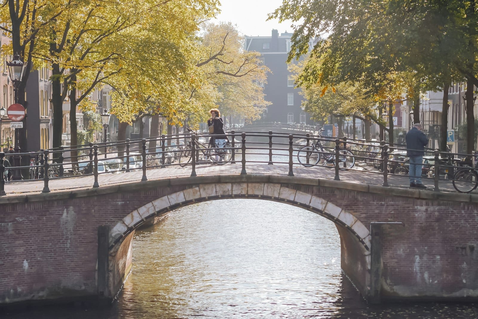 Escena de canal de Ámsterdam iluminada por el sol con un puente de piedra, personas caminando y en bicicleta, y árboles a lo largo del agua.