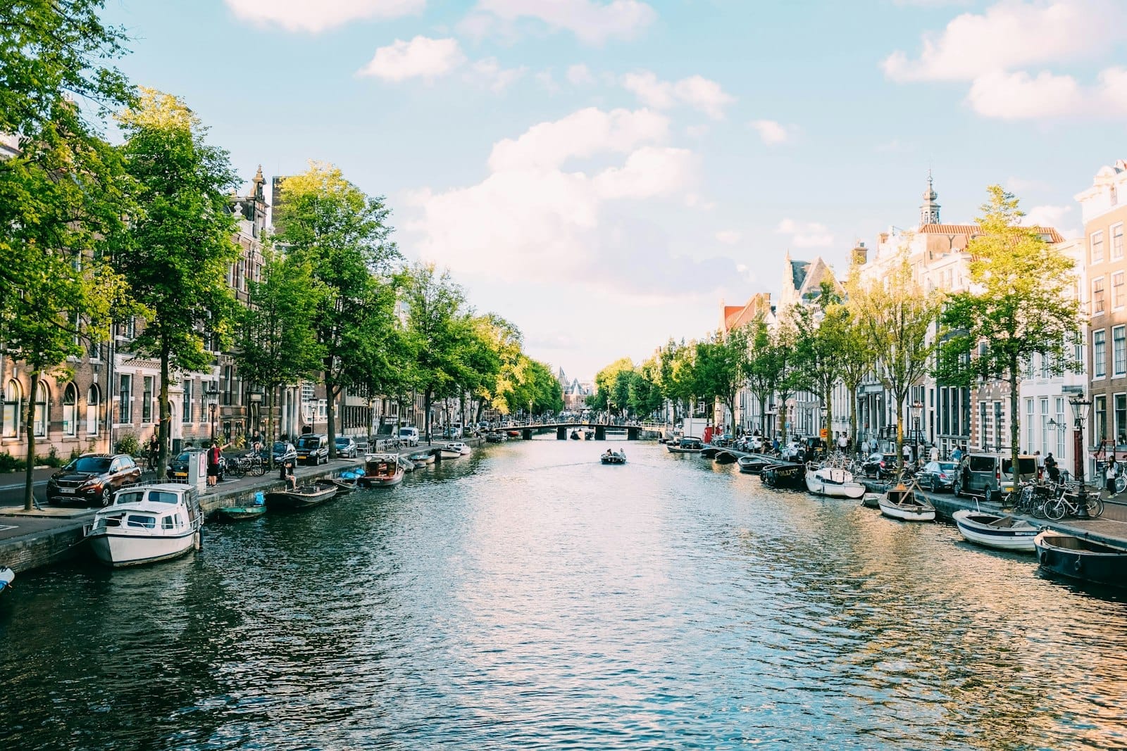 Vista soleada a lo largo de un canal de Ámsterdam bordeado de árboles, barcos y casas históricas, con personas caminando y en bicicleta a ambos lados bajo un cielo azul con nubes.