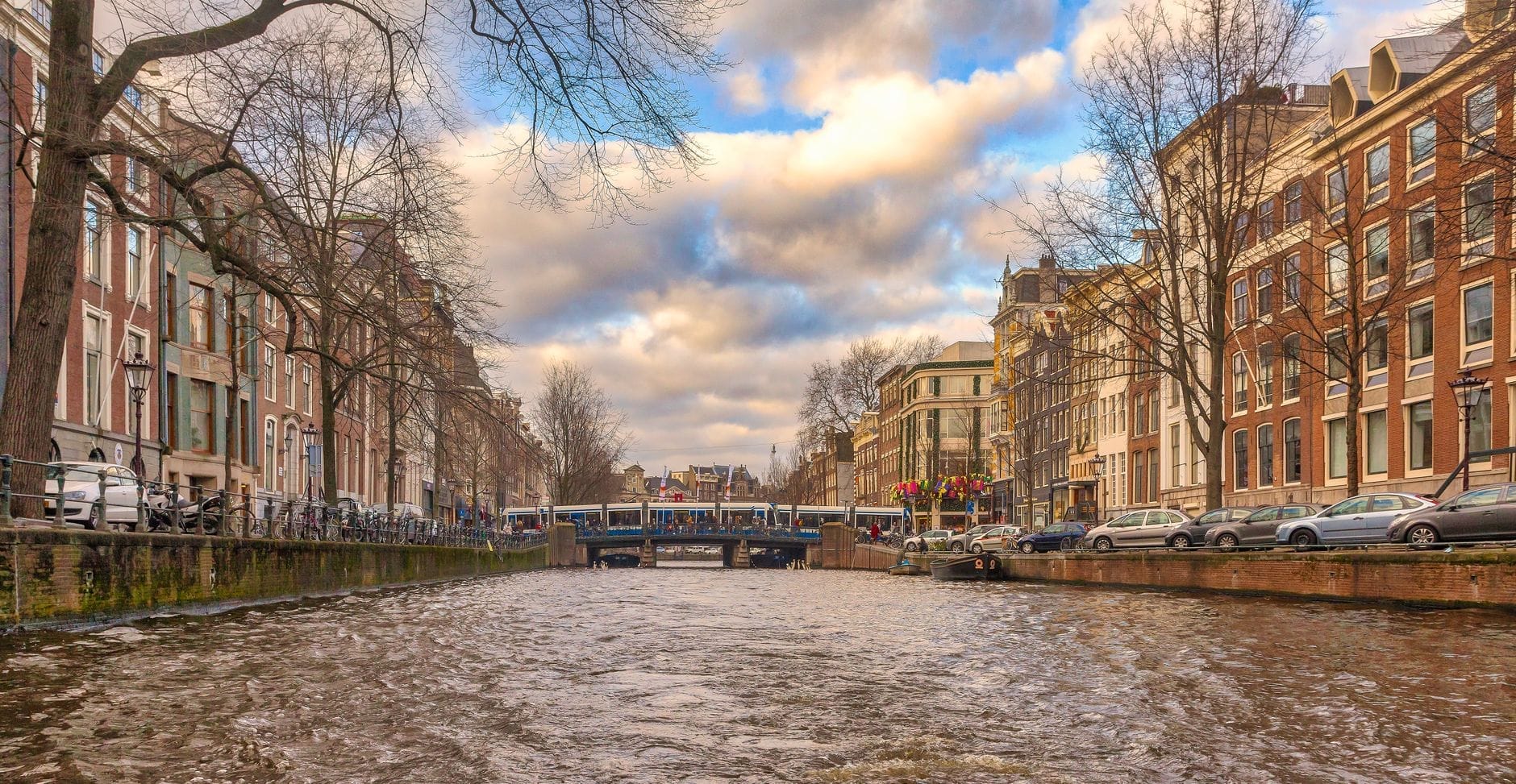Vista panorámica de un canal de Ámsterdam con casas clásicas a ambos lados, un tranvía cruzando un puente y nubes dramáticas en el cielo.