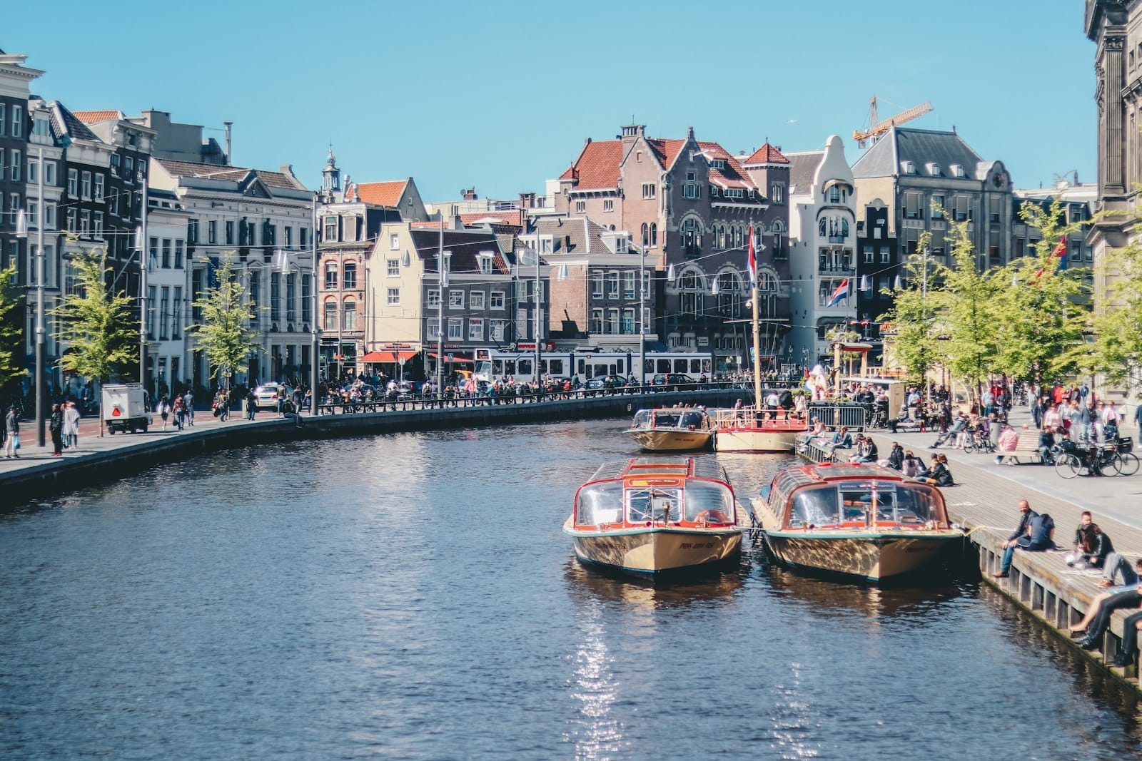 Escena de canal en Ámsterdam con barcos turísticos, edificios históricos y personas sentadas junto al agua en un día soleado.