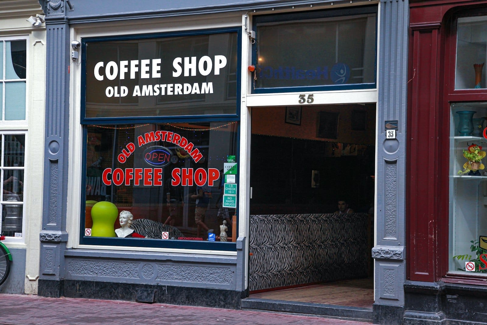 La entrada al coffee shop 'Old Amsterdam' con grandes ventanales, letreros y un vistazo al interior que muestra asientos y decoración típicos de los coffeeshops de Amsterdam.