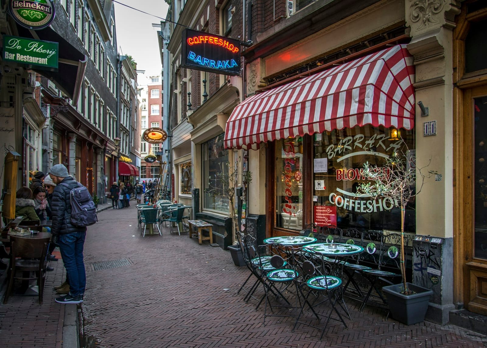 Vista de la calle de un coffeeshop clásico de Ámsterdam con mesas al aire libre, letreros de neón y personas caminando por un animado callejón estrecho.