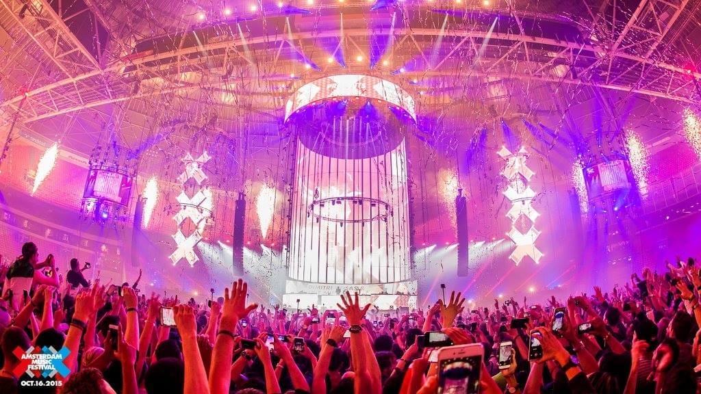Una gran multitud baila bajo luces de escenario vibrantes y decoraciones del Amsterdam Dance Event dentro de un recinto musical abarrotado durante un concierto en vivo.
