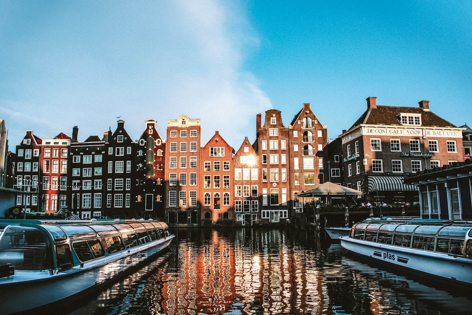 Escena clásica de canal en Ámsterdam con casas holandesas tradicionales y dos barcos de crucero fluvial con techo de cristal al atardecer.