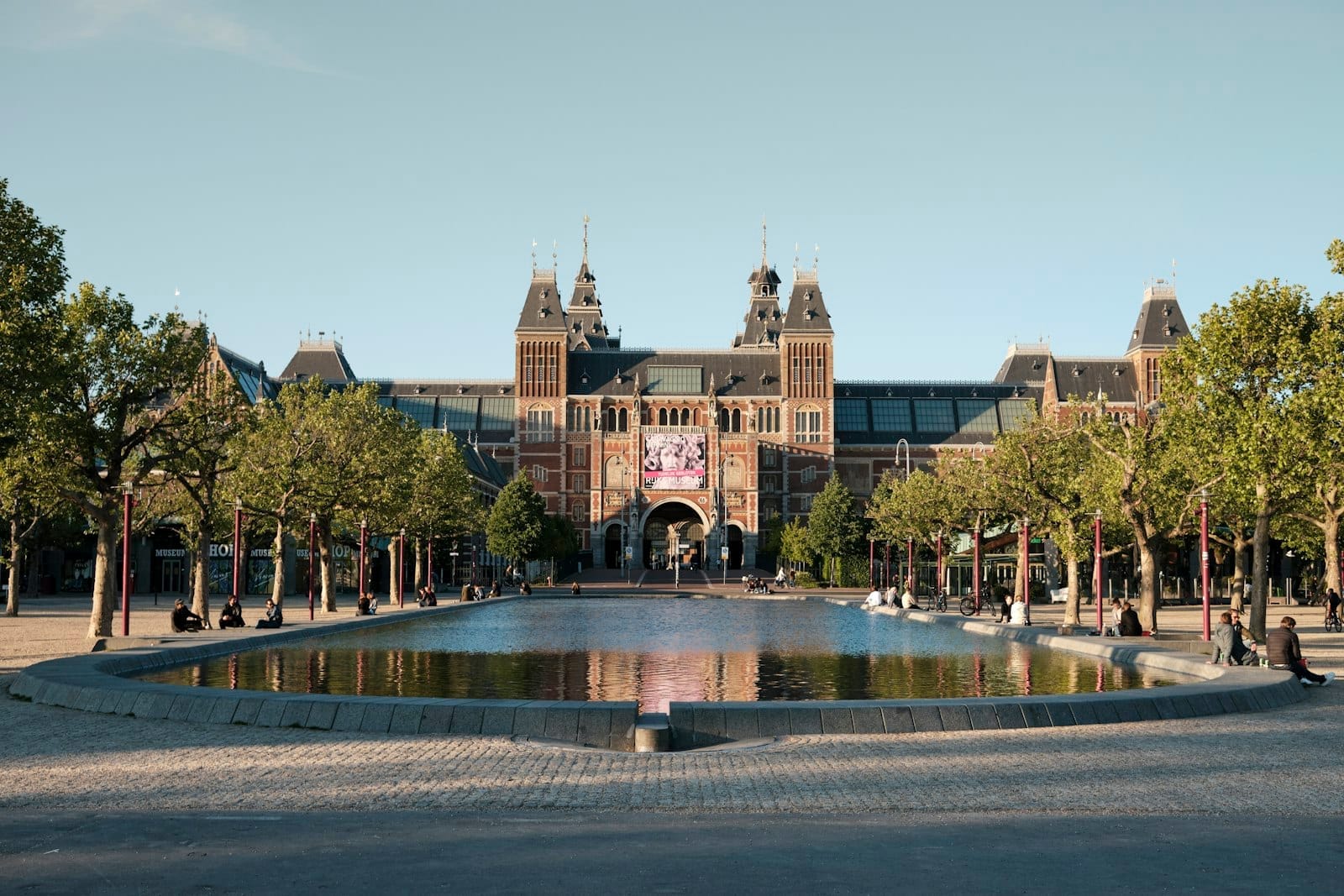 La fachada del Rijksmuseum en Ámsterdam, enmarcada por árboles y reflejada en una gran fuente en un día soleado.