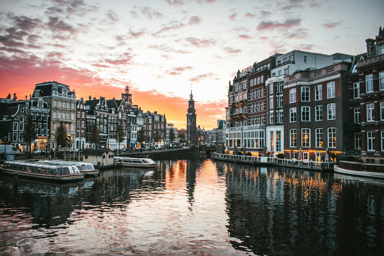 Vista romántica al atardecer de un canal de Ámsterdam flanqueado por edificios históricos, barcos y reflejos luminosos en el agua, creando una atmósfera perfecta para parejas.