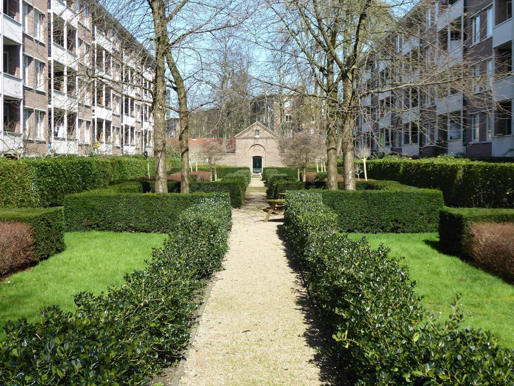 Jardín verde de patio flanqueado por edificios de apartamentos estilo canal en Amsterdam, que conduce a la pequeña entrada de una capilla de ladrillo.