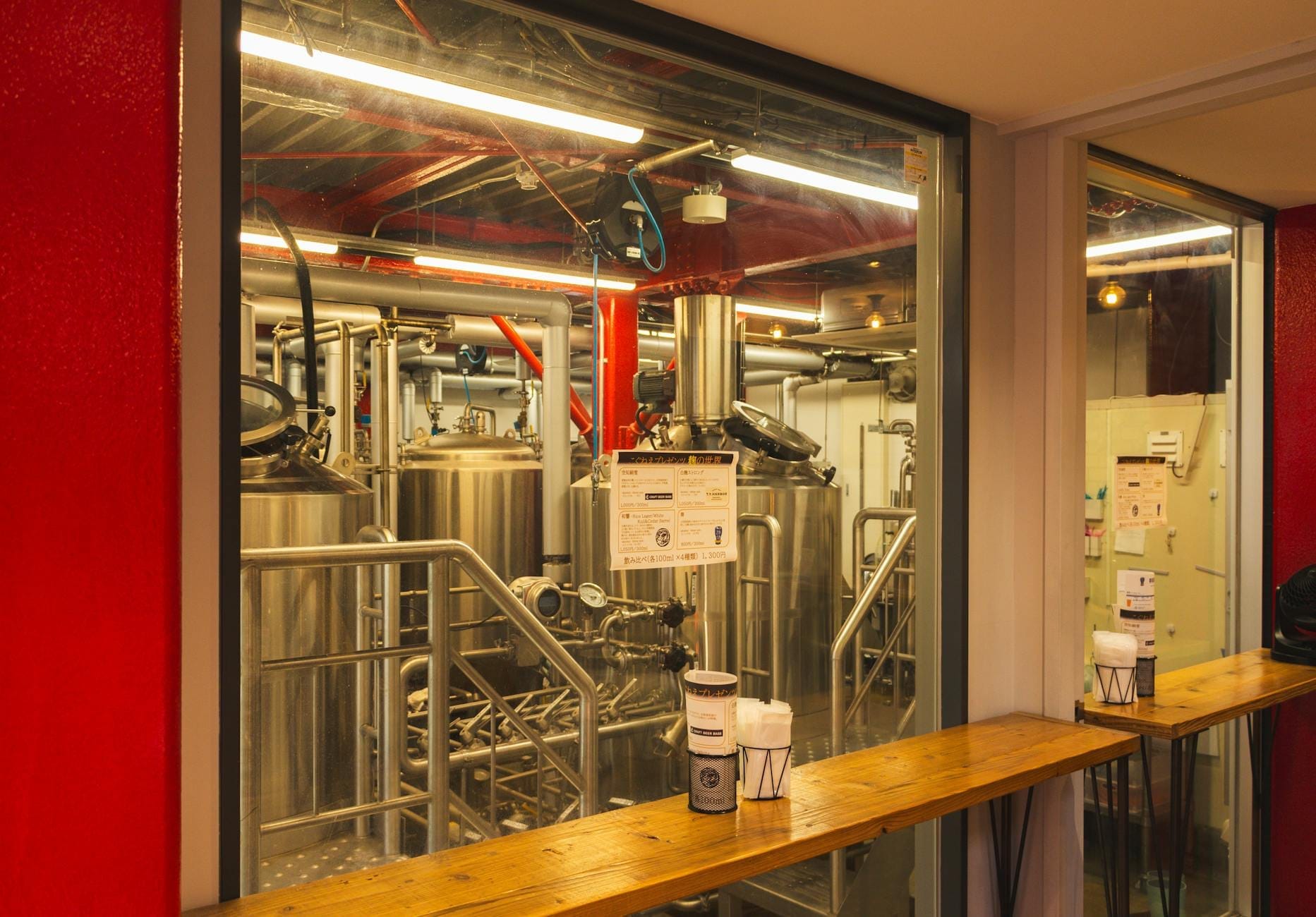 Vista interior de una cervecería artesanal con equipos de elaboración visibles detrás de un cristal, ubicada en un espacio industrial moderno con paredes rojas y asientos en barra.