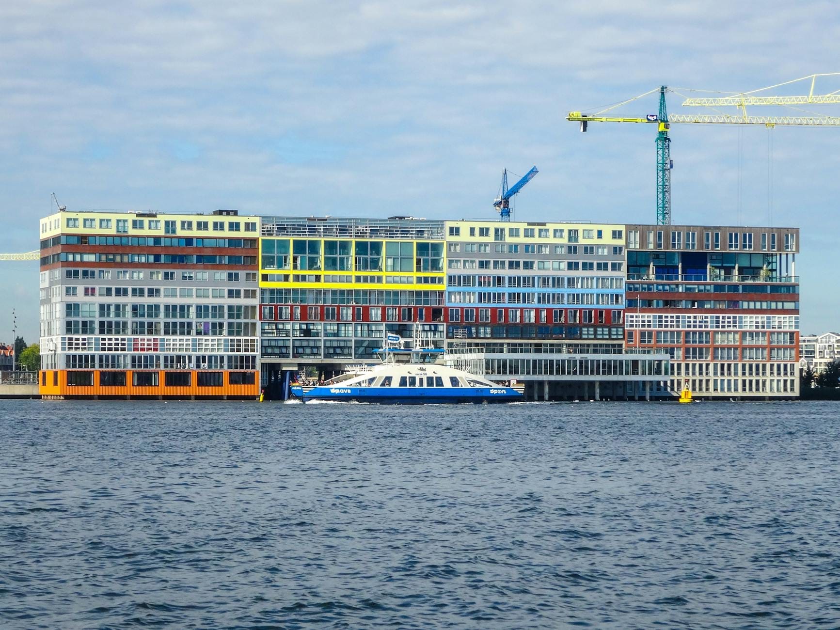 Edificio moderno en Amsterdam-Noord visto desde el otro lado del IJ, con un ferry y grúas de construcción visibles.