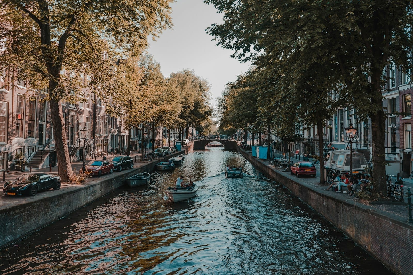 Un tranquilo canal de Ámsterdam enmarcado por frondosos árboles y casas tradicionales, con pequeñas embarcaciones navegando y gente relajándose junto al agua bajo una suave luz diurna.
