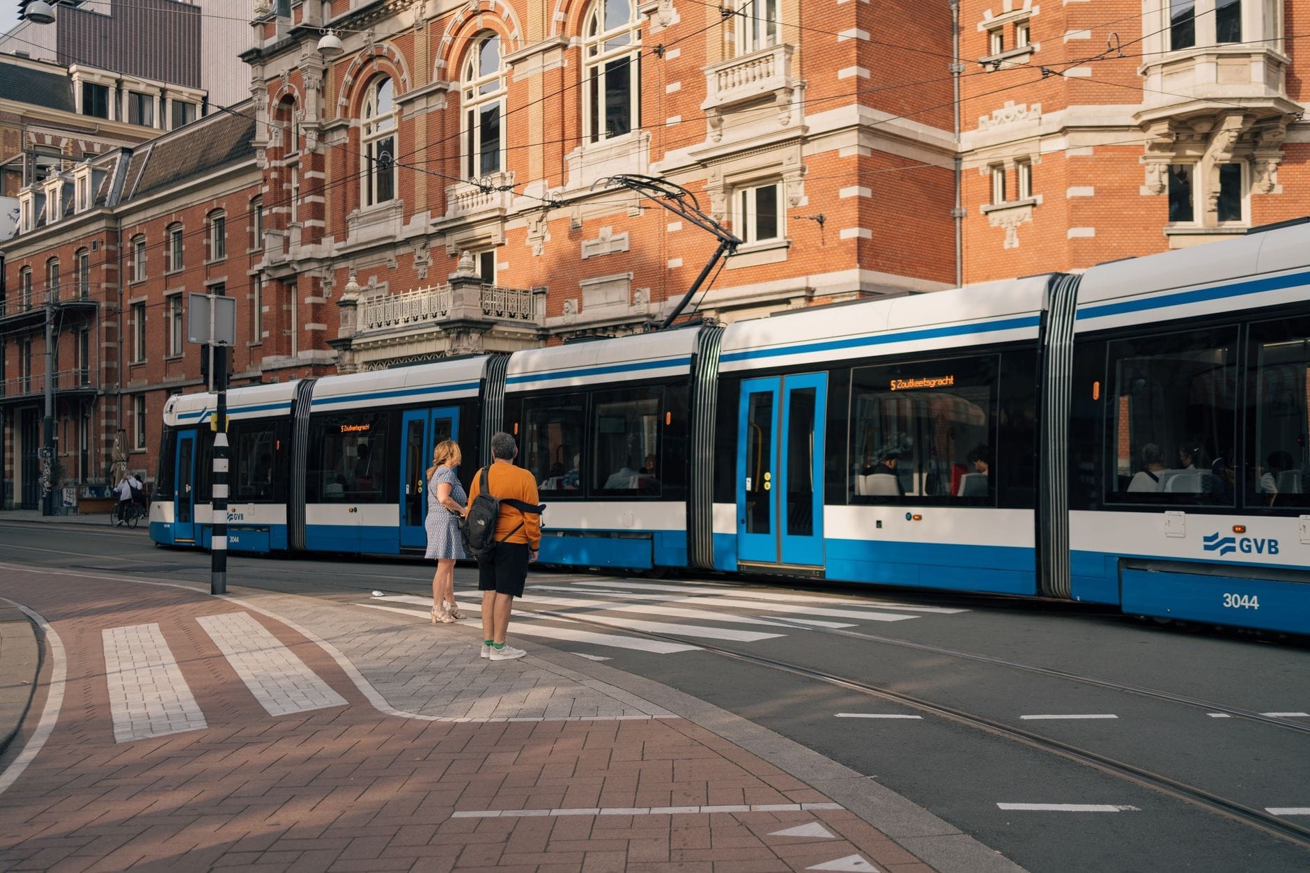 Tranvía moderno azul y blanco de GVB con personas esperando en una parada frente a la arquitectura tradicional de Amsterdam.