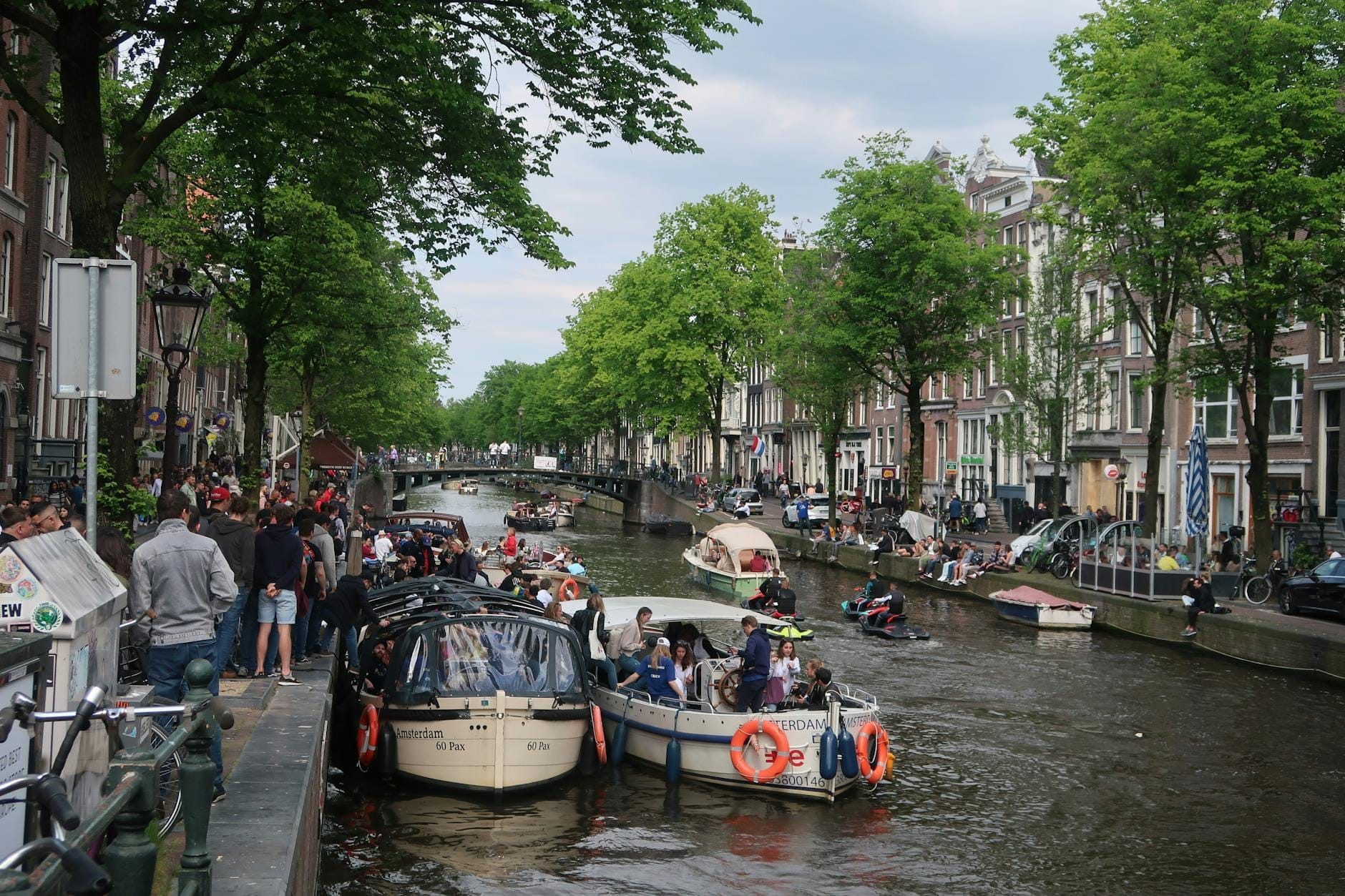 Canal de Amsterdam lleno de barcos y personas a lo largo de ambas orillas disfrutando de festividades, animado con grupos y decoraciones naranjas.