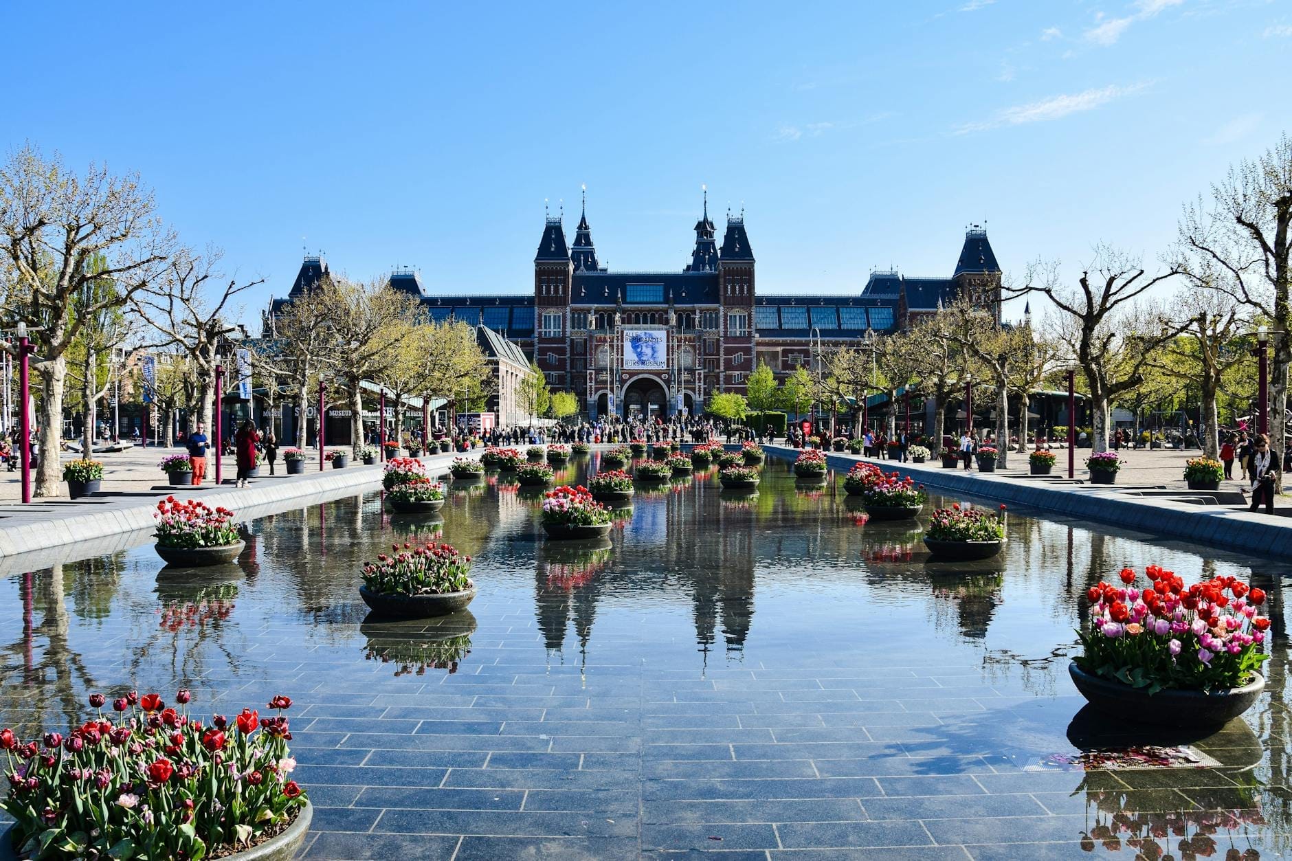Vista del Rijksmuseum en Ámsterdam con un estanque reflectante rodeado de tulipanes en flor y árboles bajo un cielo azul despejado de primavera.