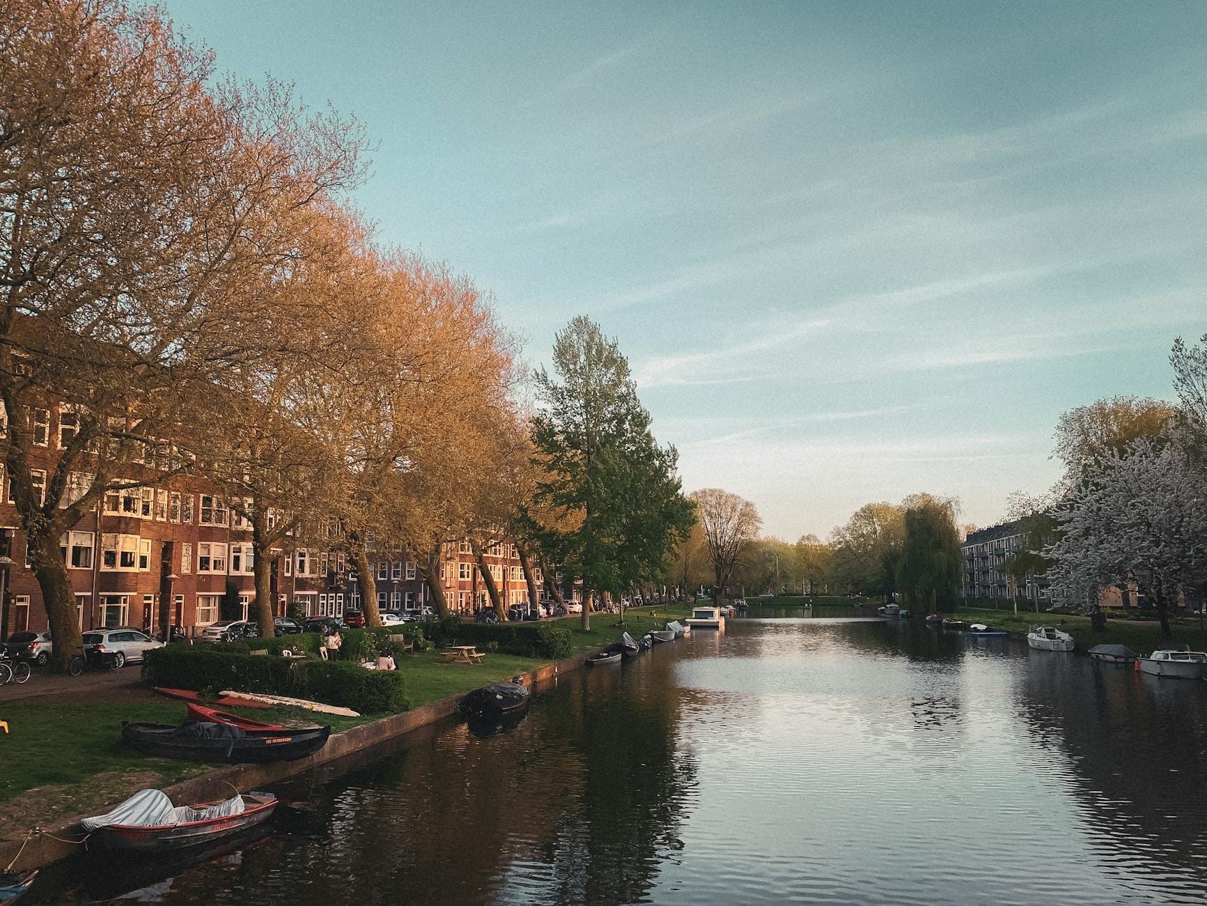 Canal tranquilo de Ámsterdam en primavera bordeado de árboles en flor, barcos amarrados y edificios holandeses clásicos bajo un cielo azul.